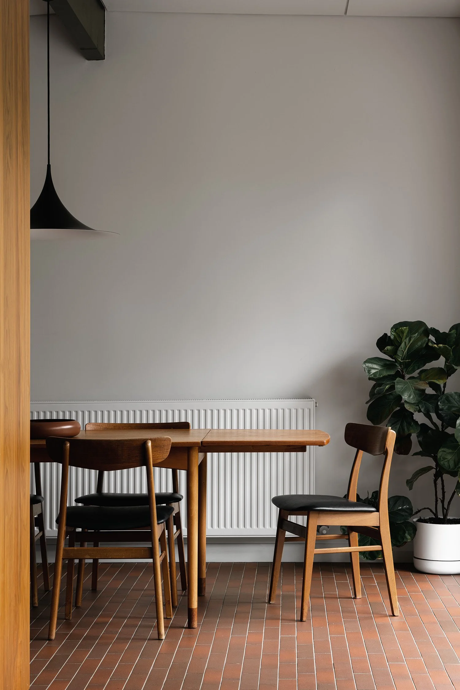 Mid century dining area with timber chairs, a black pendant light and Japanese terracotta floor tiles beside a leafy indoor plant.