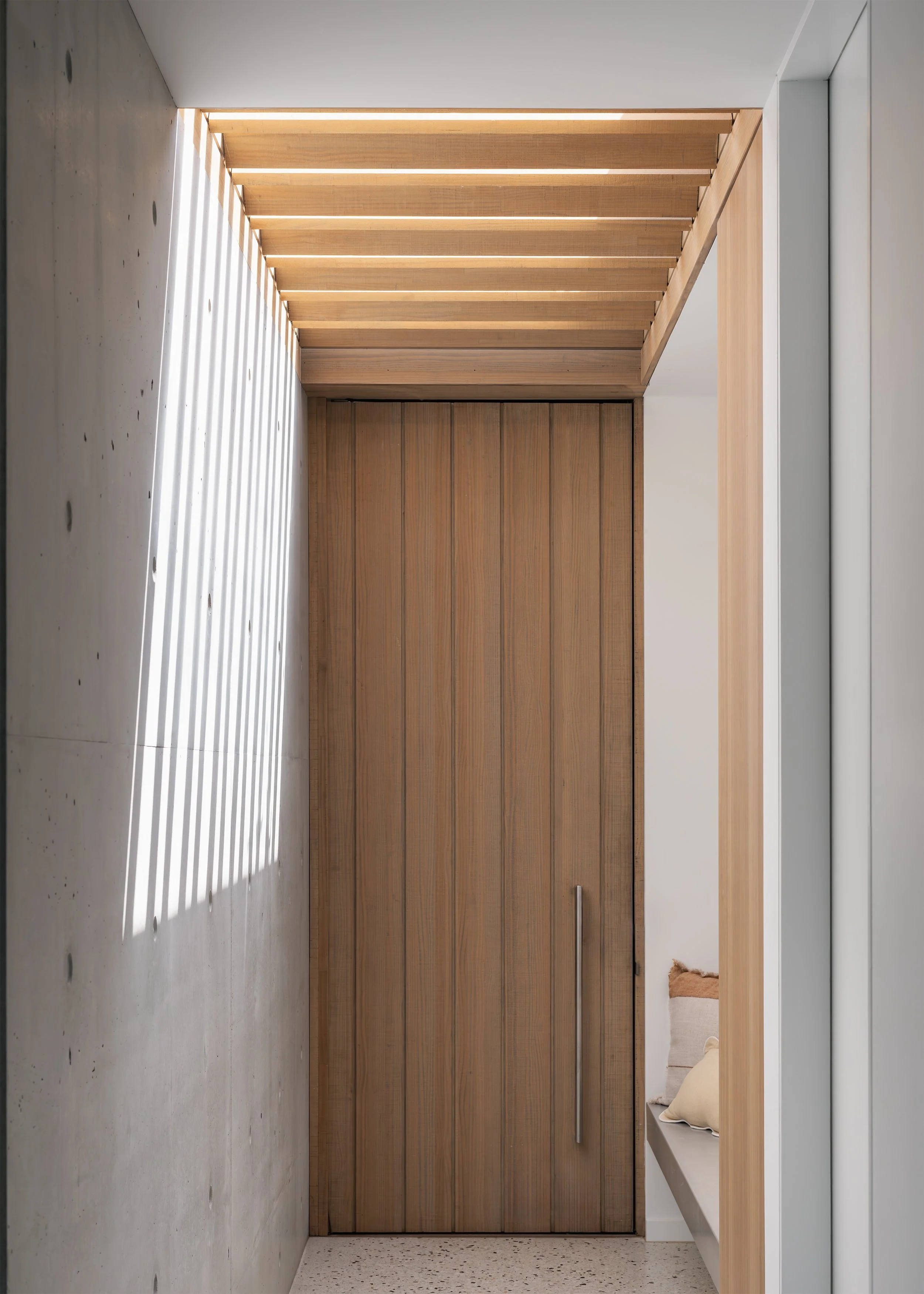 Timber front door framed by exposed concrete, timber battens and filtered light at Channel in Mornington.
