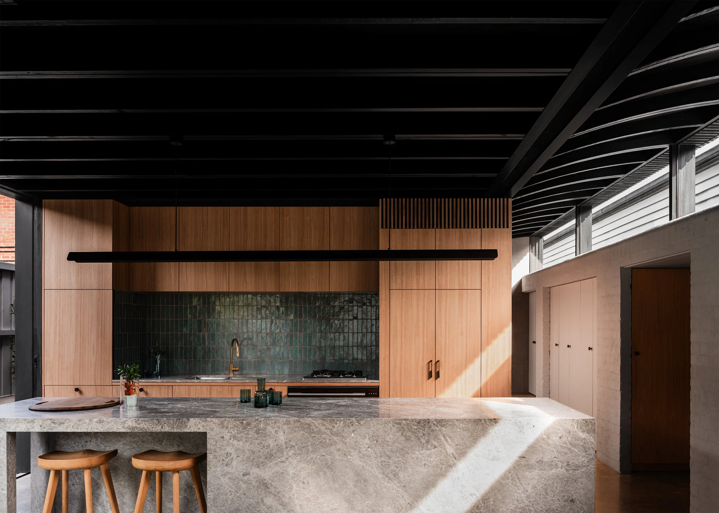 Kitchen joinery and stone island lit by afternoon sun beneath the dark timber ceiling.