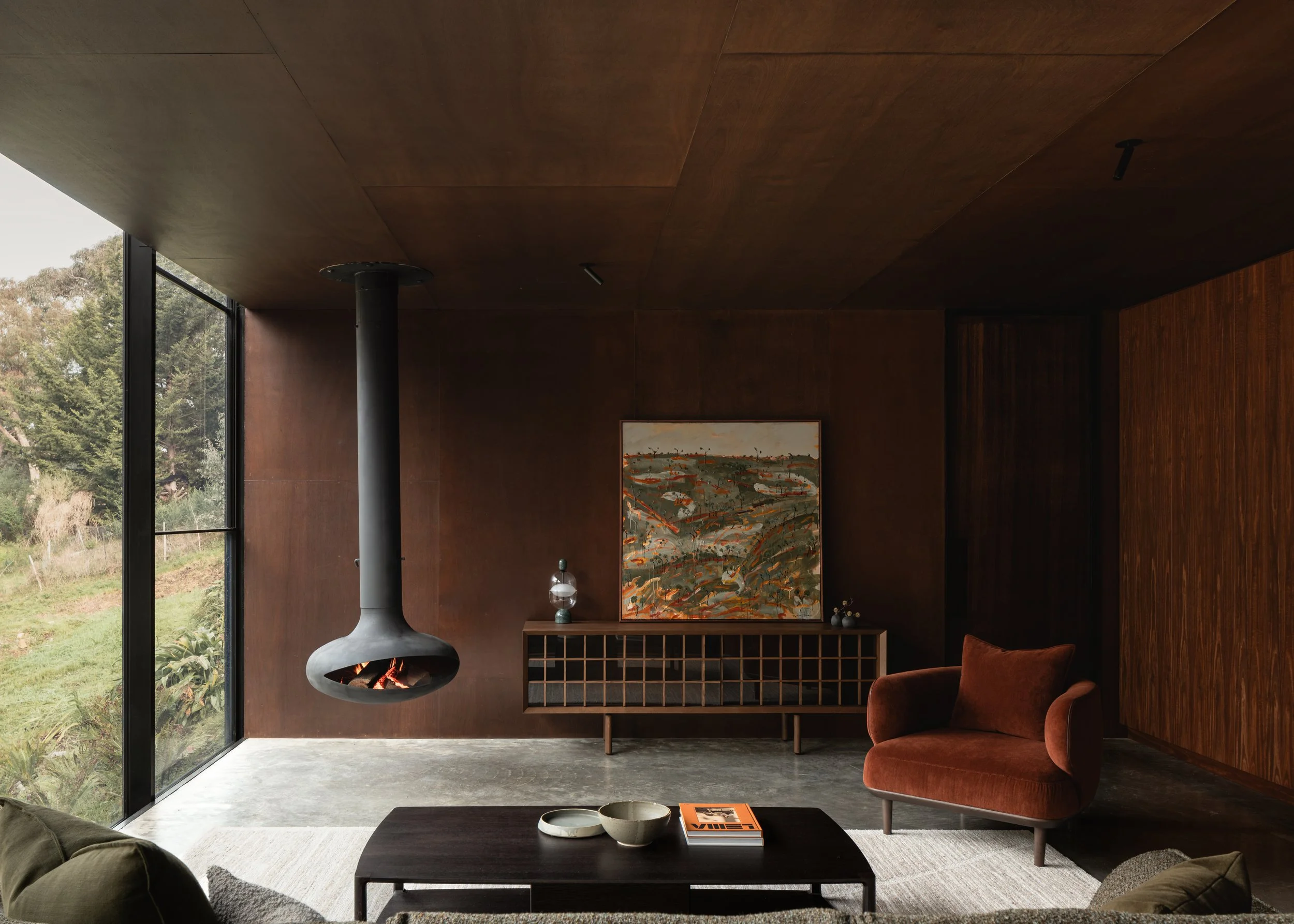Living room at Matchbox with dark timber linings, suspended fireplace and full-height glazing to the bush.