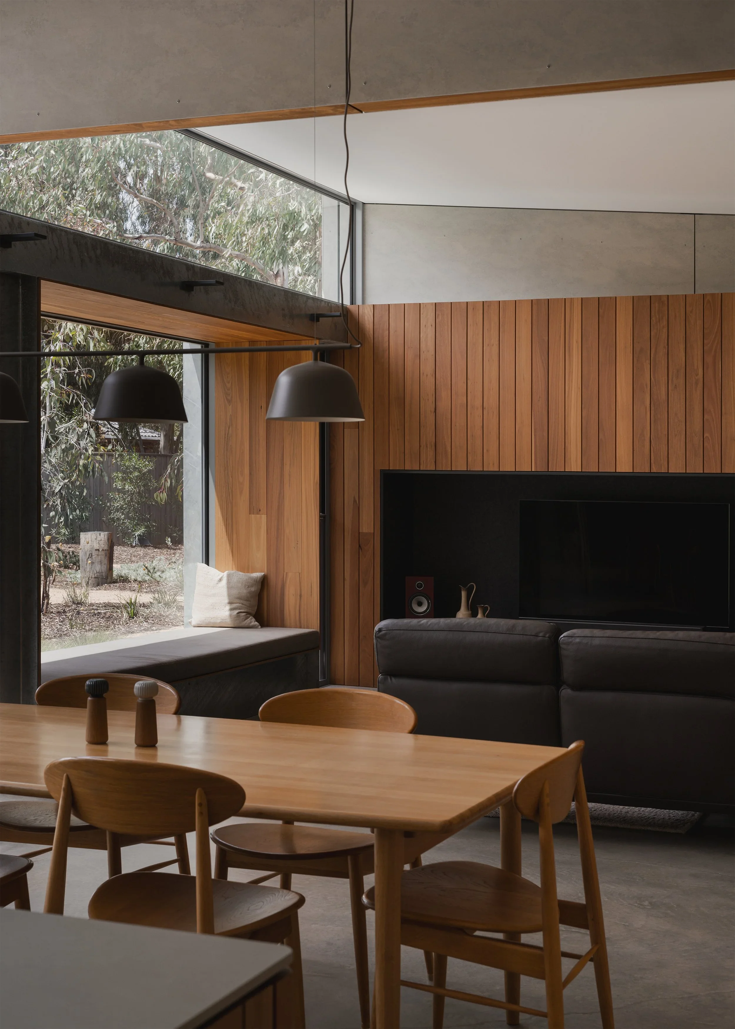 Timber dining table beneath black pendant lights at Forest Road House, with a window seat, leather sofa and timber-panelled wall beyond.