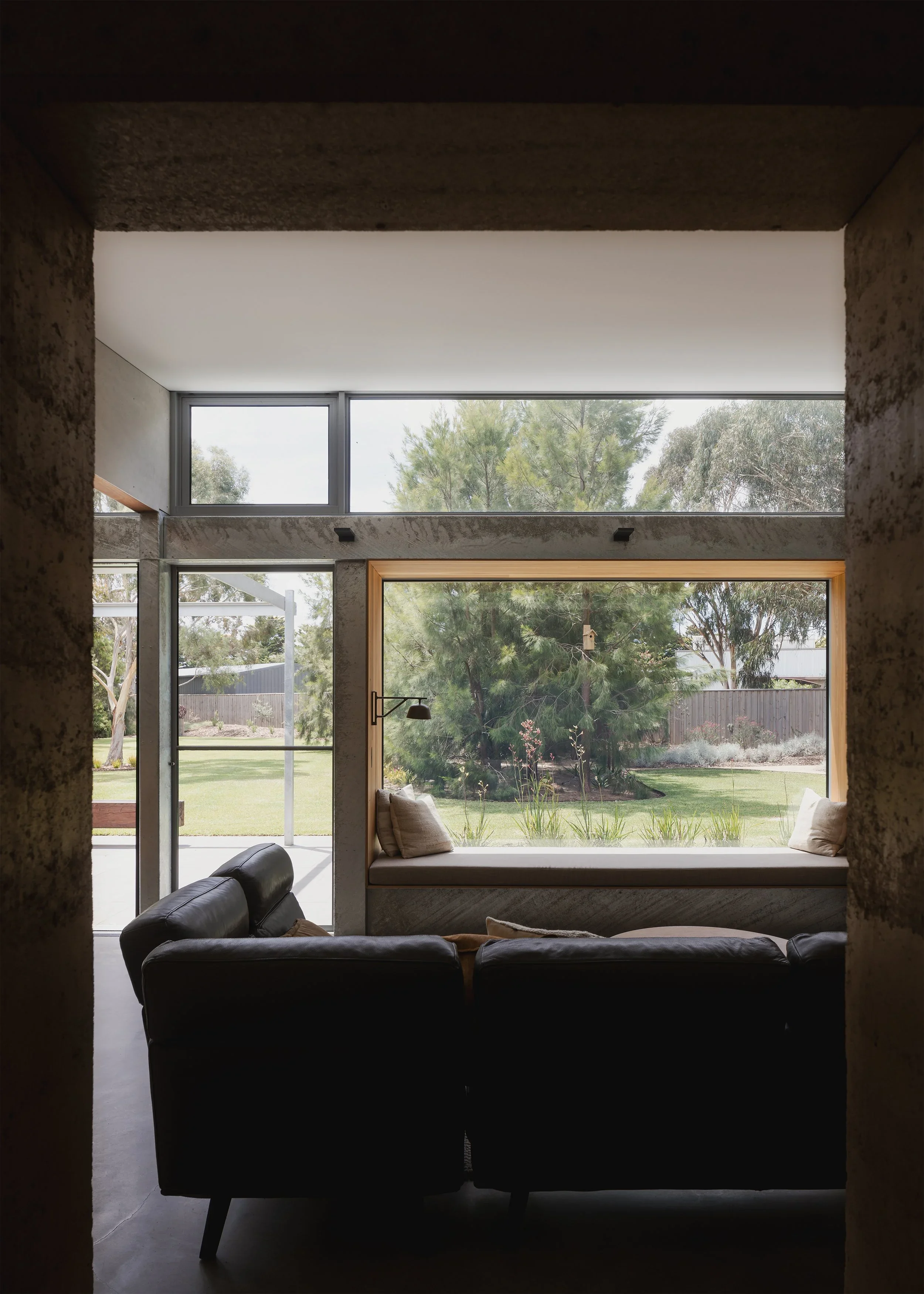 View through a rammed-earth opening into Forest Road House’s living room, with a timber-lined window seat overlooking the garden.