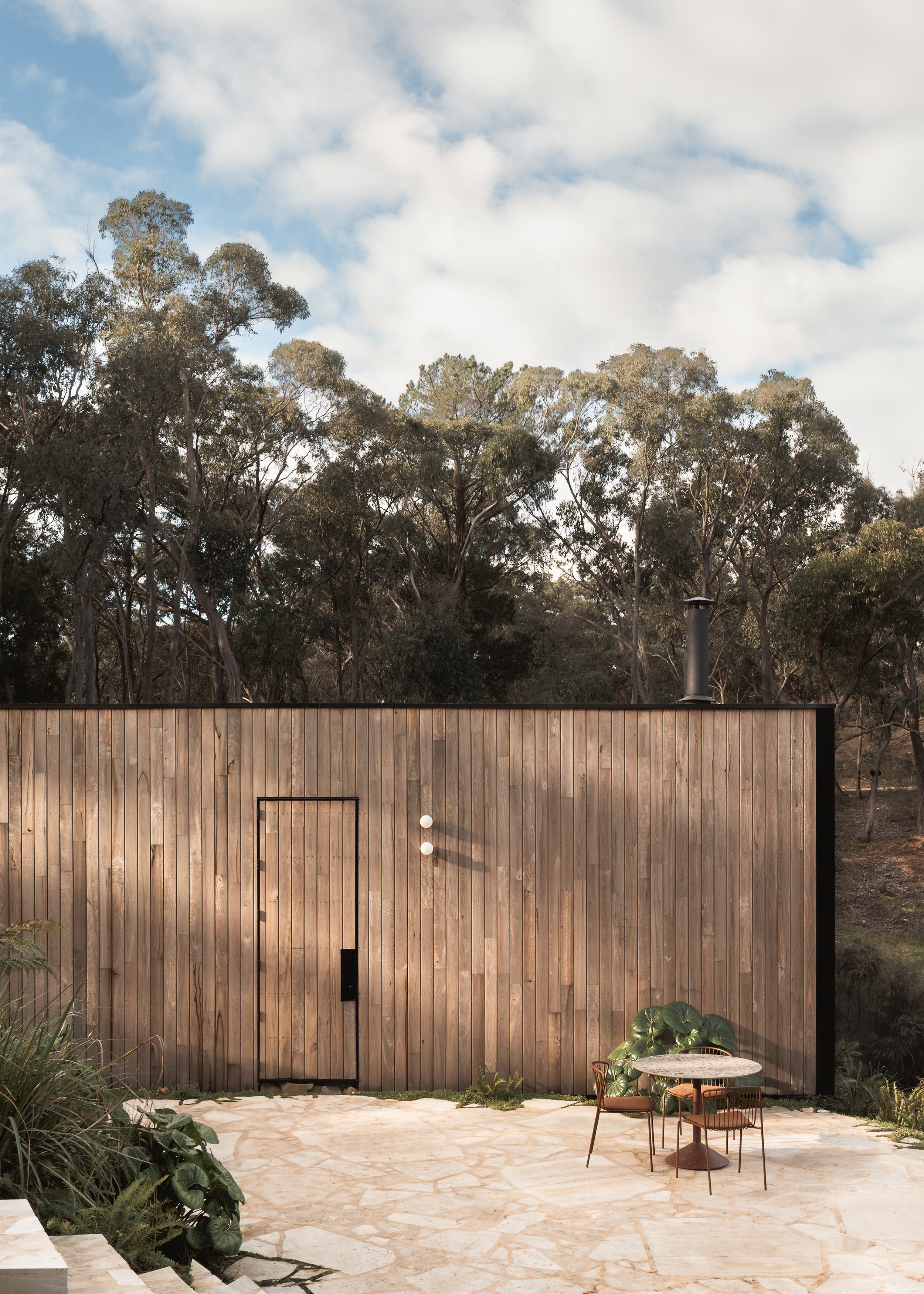 Timber-clad entrance elevation of Matchbox opening onto a crazy paving terrace beneath the surrounding eucalypts.
