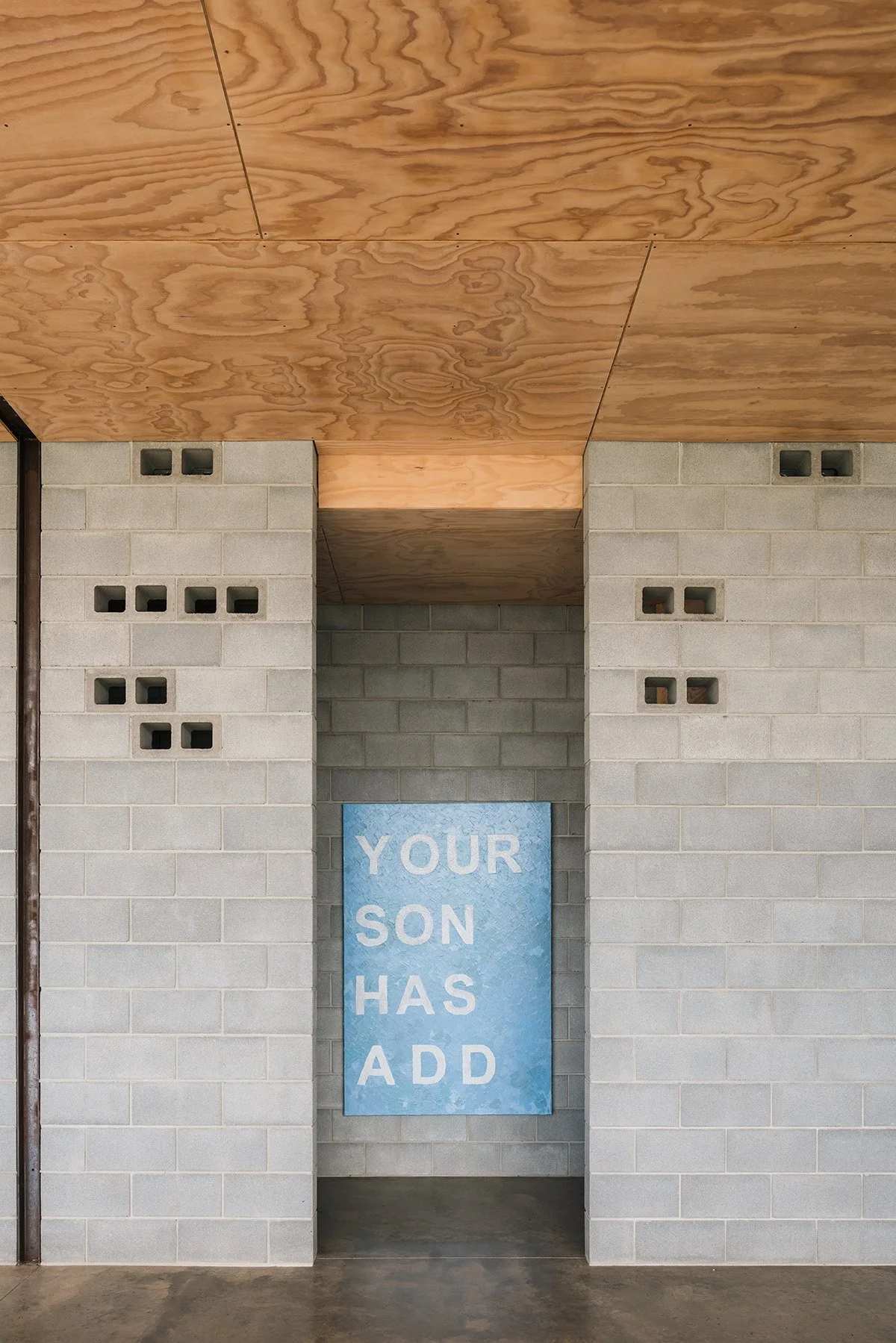 Minimalist passage at Mansfield House with concrete block walls, a plywood ceiling, breeze-block openings and blue text artwork.