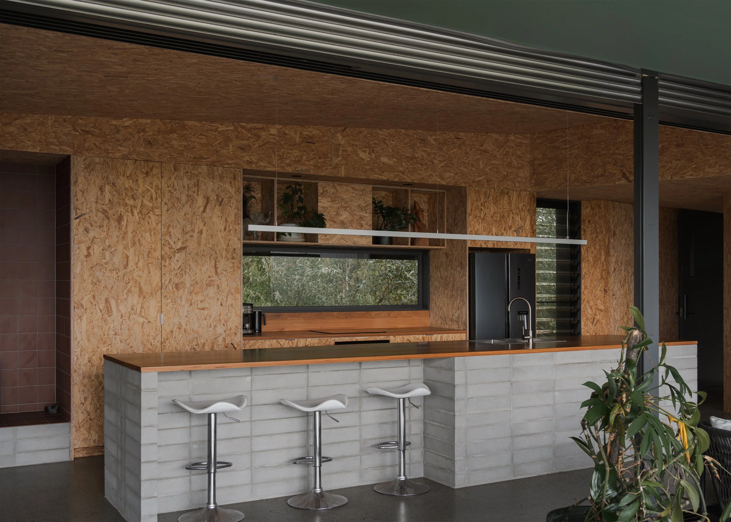 Open kitchen at Tallowwood Cabin with a concrete-block island, timber benchtop, OSB joinery and a long window framing the tree canopy.