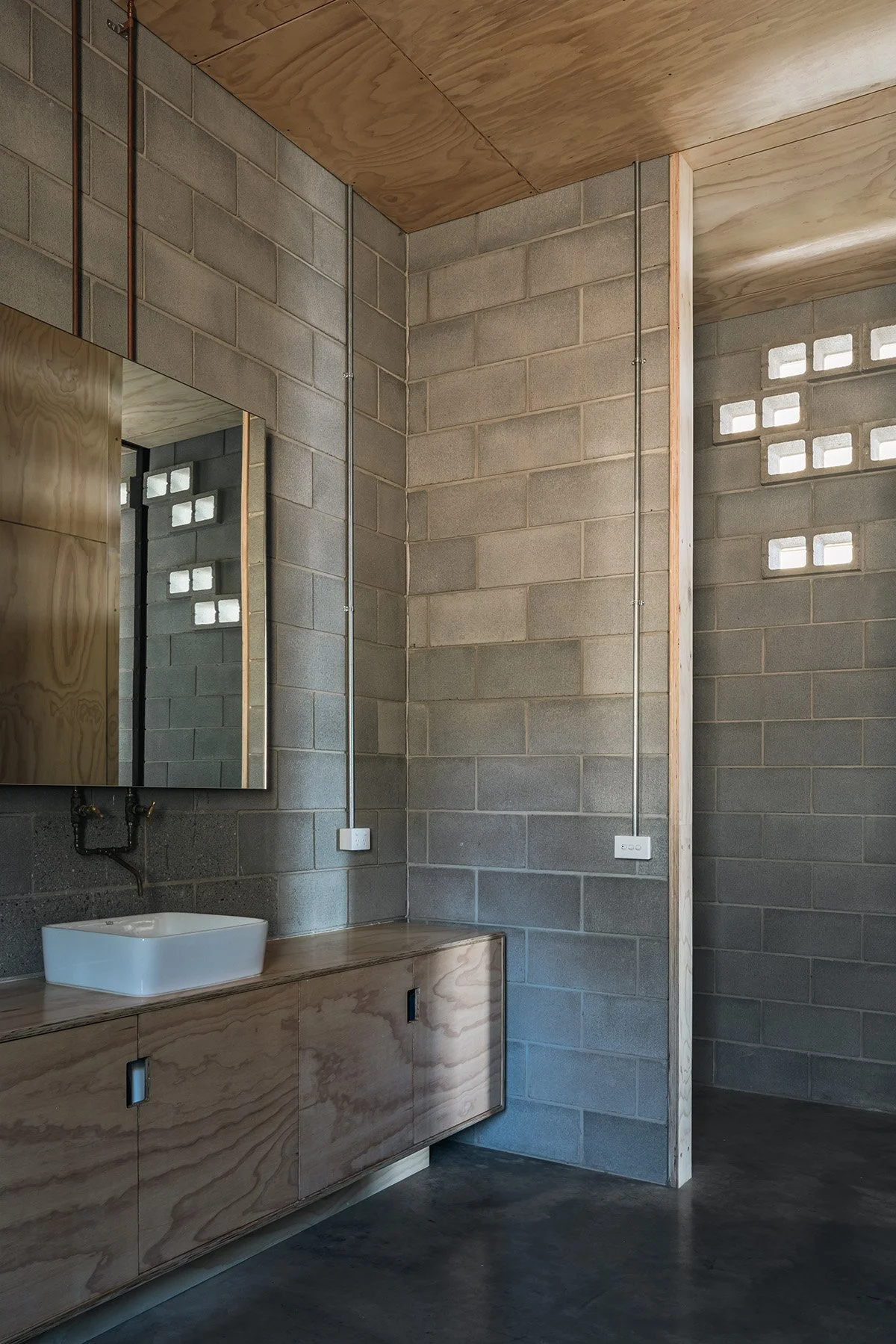 Minimalist bathroom with concrete block walls, a plywood vanity and ceiling, and patterned breeze-block openings.