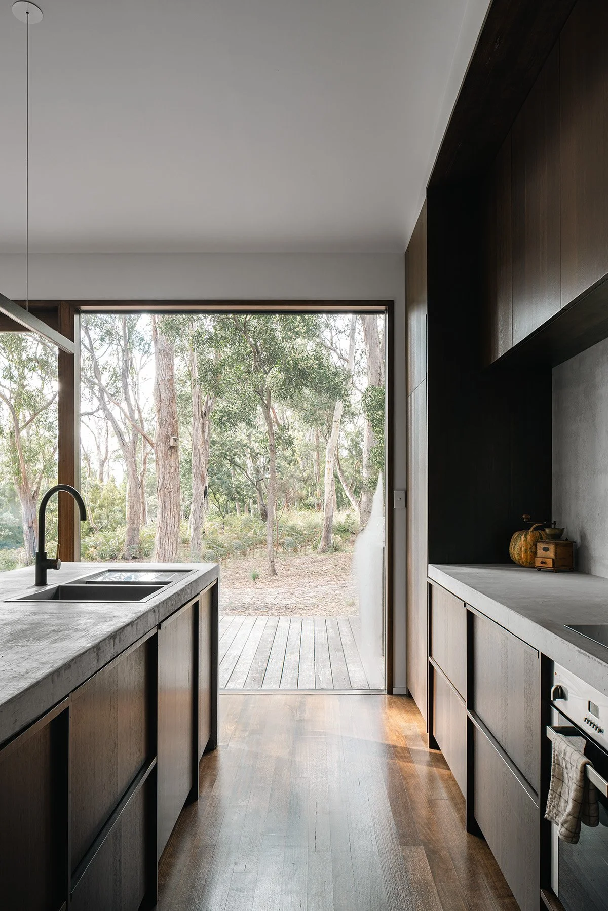 Galley kitchen at Killora Bay with concrete benchtops, dark timber cabinetry and a direct outlook to the Bruny Island bush.