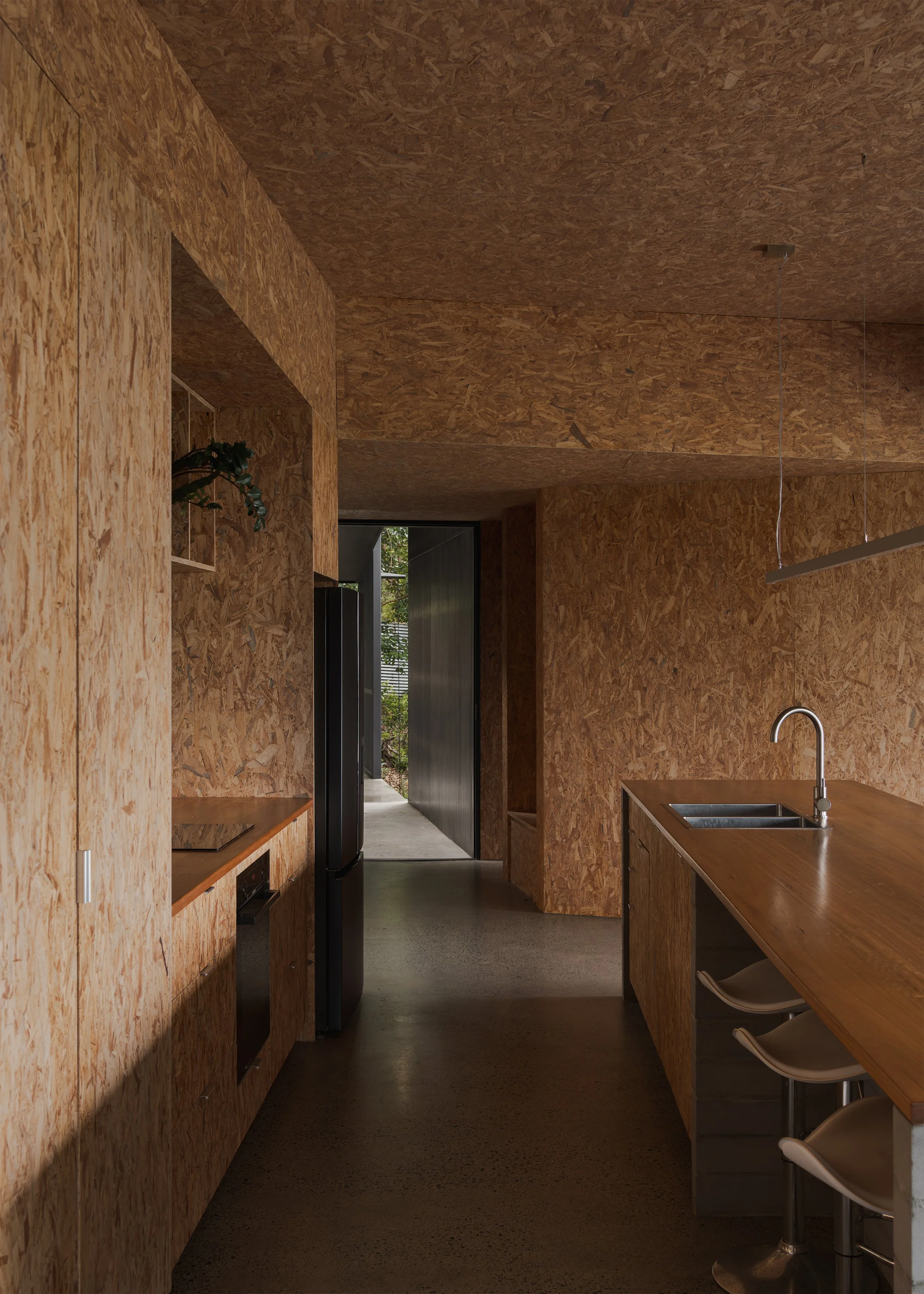 Galley kitchen at Tallowwood Cabin lined in exposed OSB, with timber benchtops, polished concrete floors and a framed view to the entry.