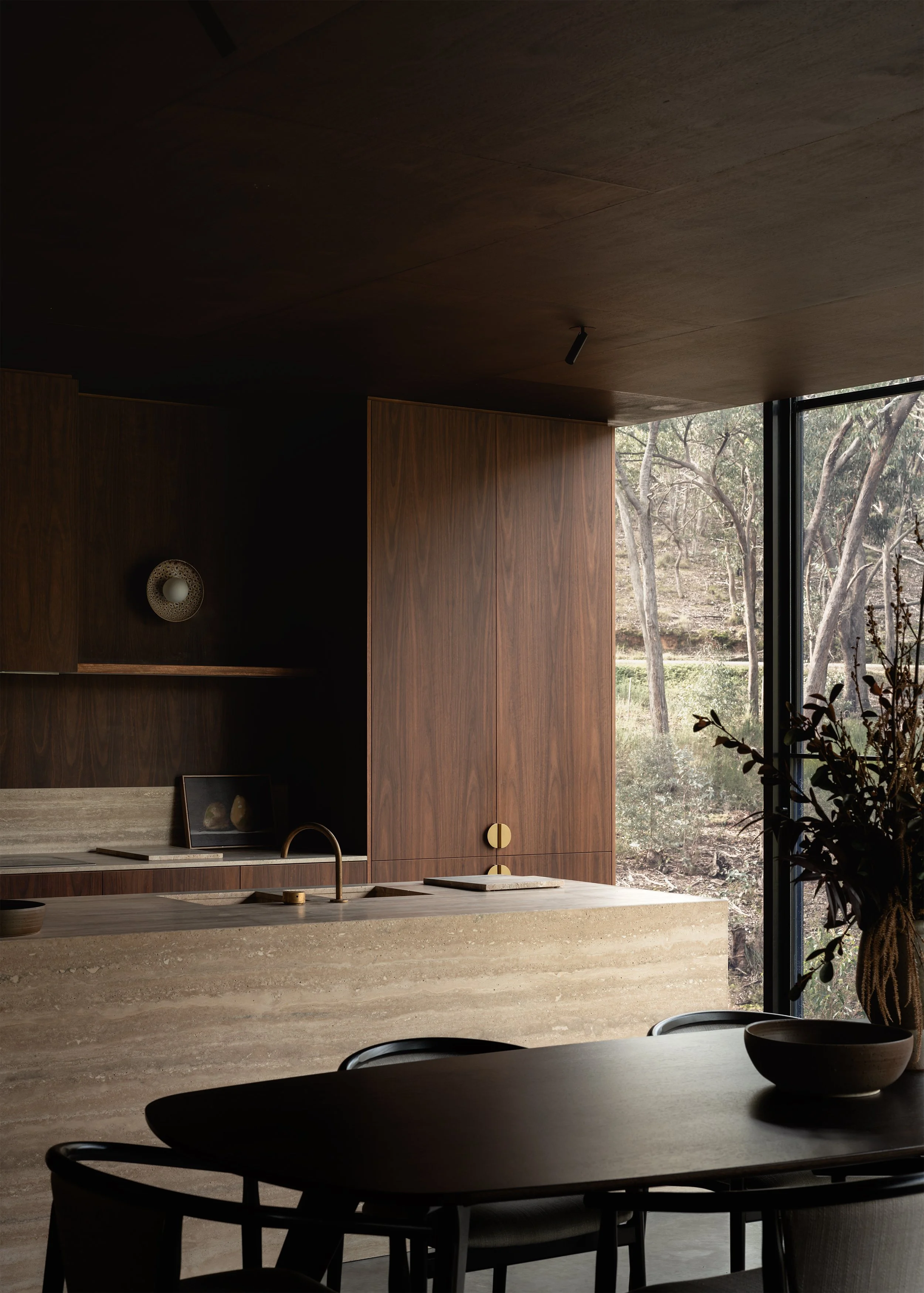 Dark timber kitchen and dining area with a travertine stone island and full-height window to the bush.
