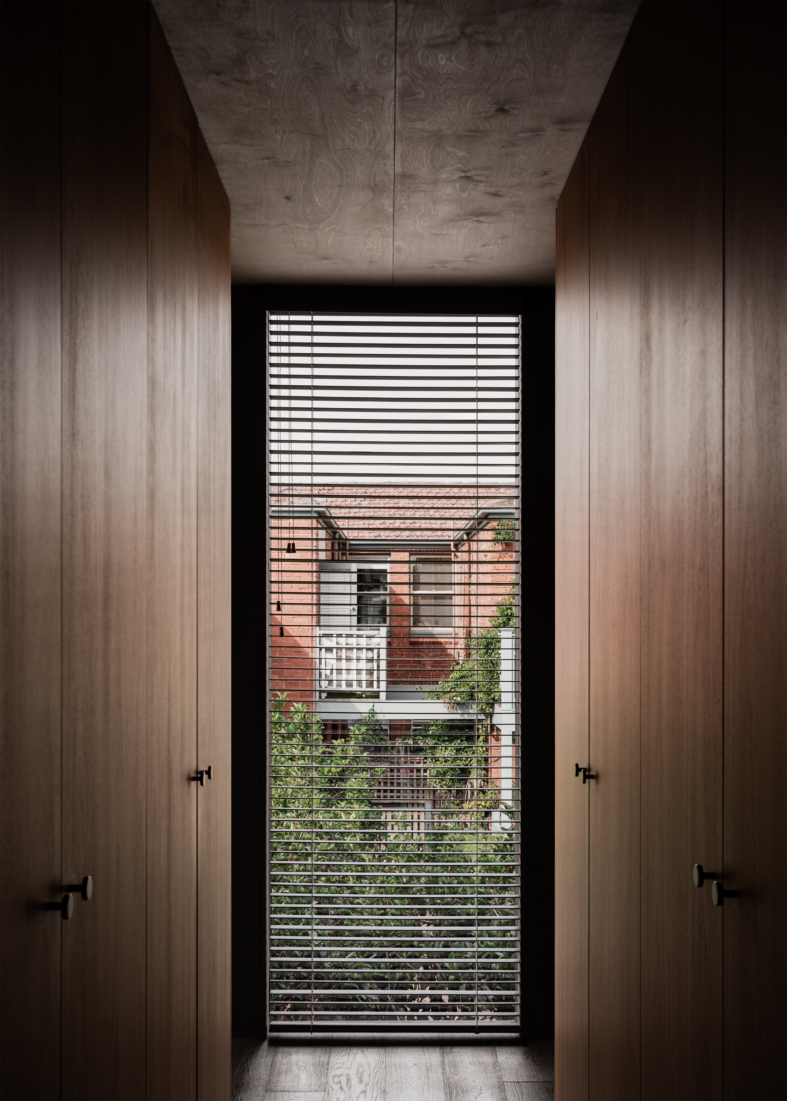 Walk-in-robe with tall window with venetian blinds framing neighbouring Victorian houses and garden from the upper level.