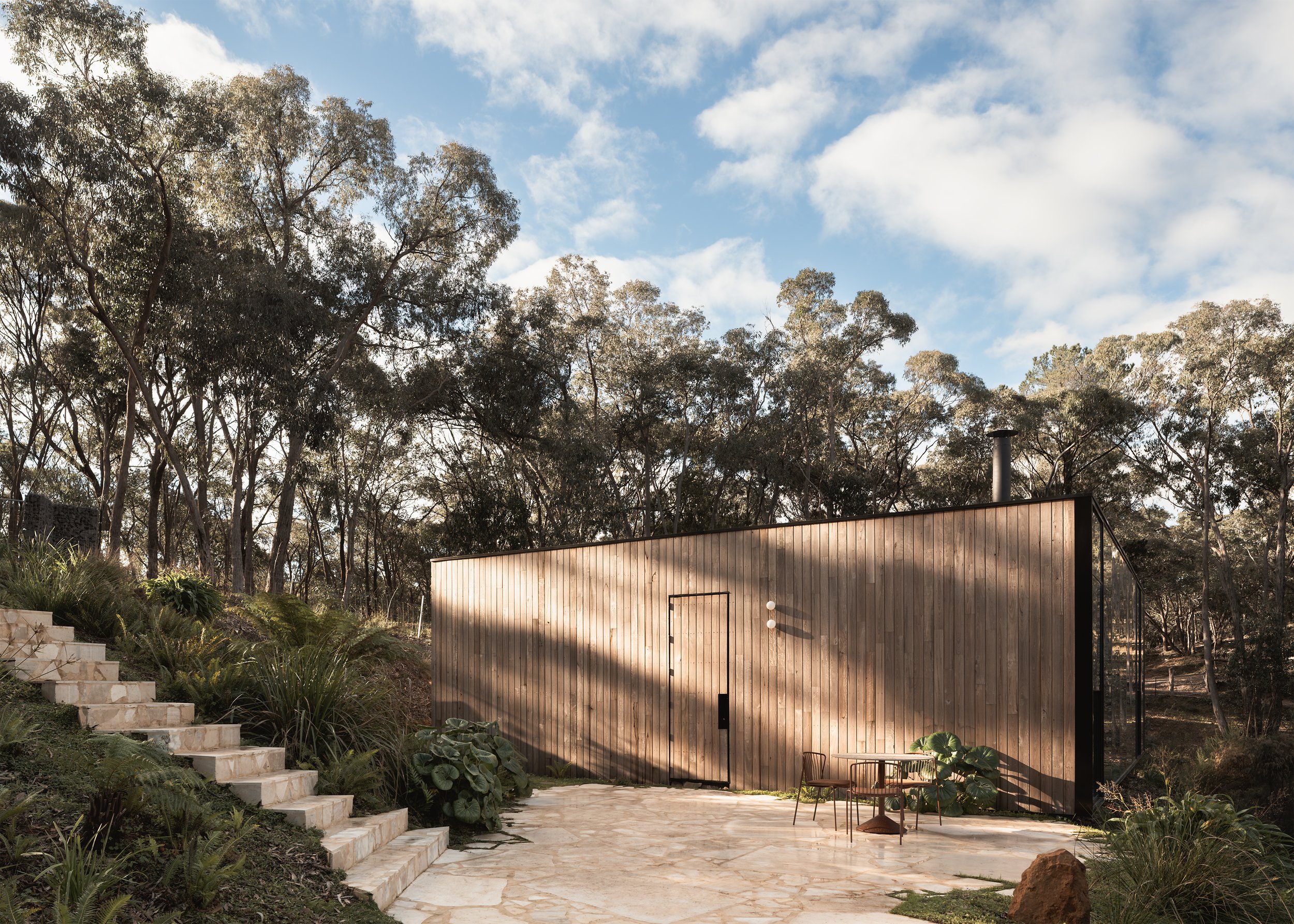 Wide exterior view of Matchbox by Vincent Jarvis Studio in Hepburn Springs with its timber facade, stone terrace, garden steps and bushland setting.