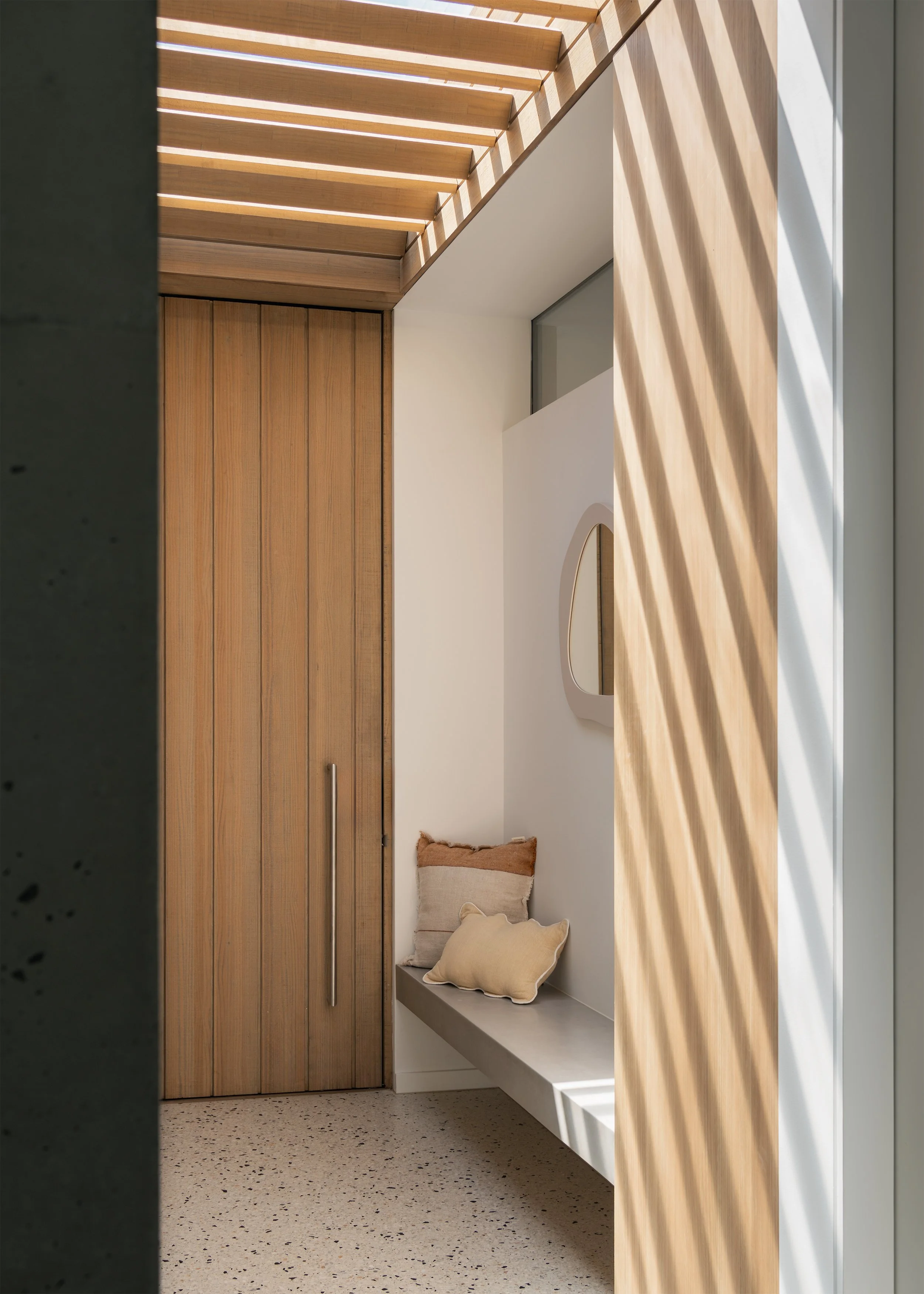 Built-in entry bench and timber door beneath a slatted timber roof casting patterned sunlight into the interior.