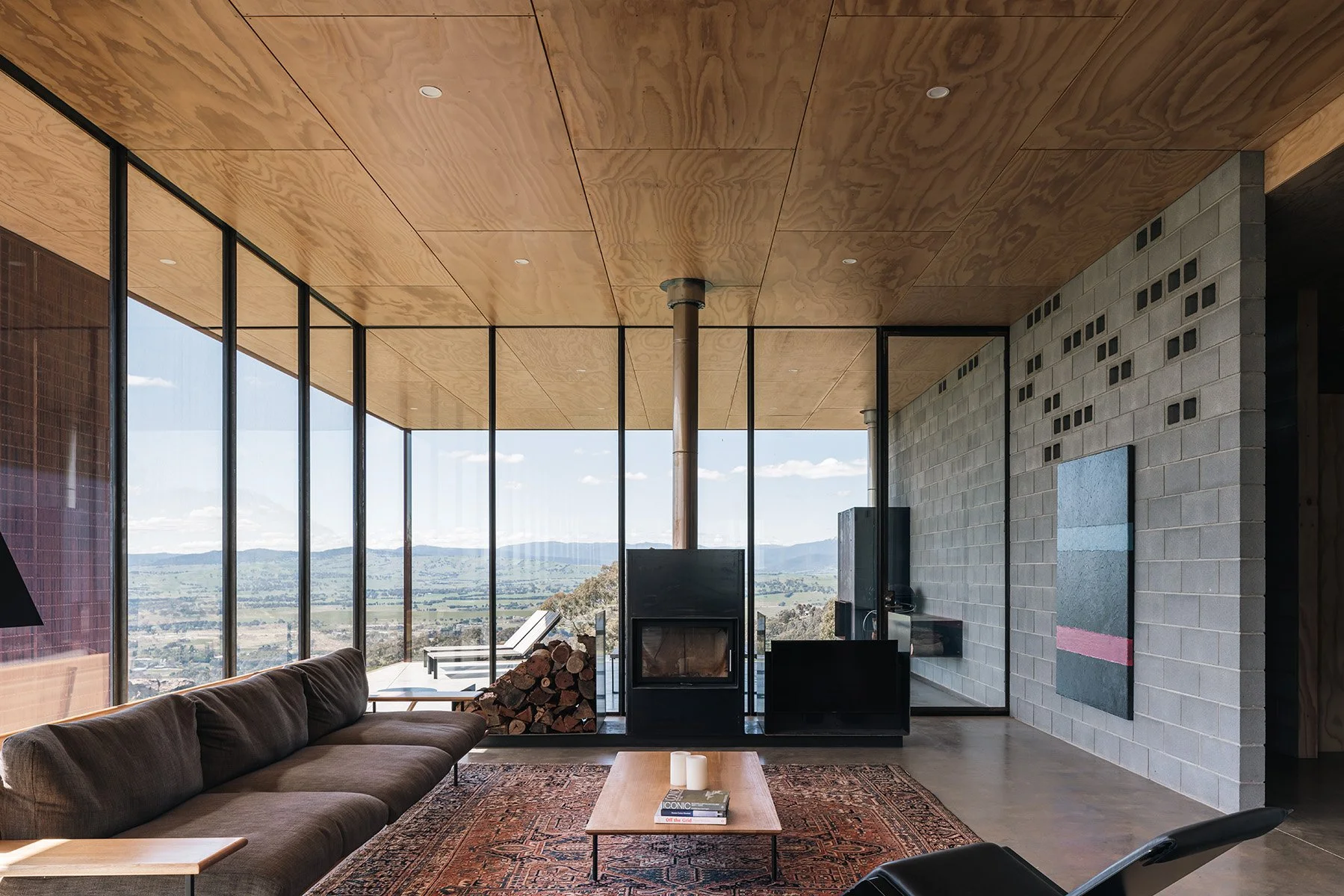 Minimalist living room at Mansfield House with concrete block walls, plywood ceiling, wood fireplace and panoramic rural views.