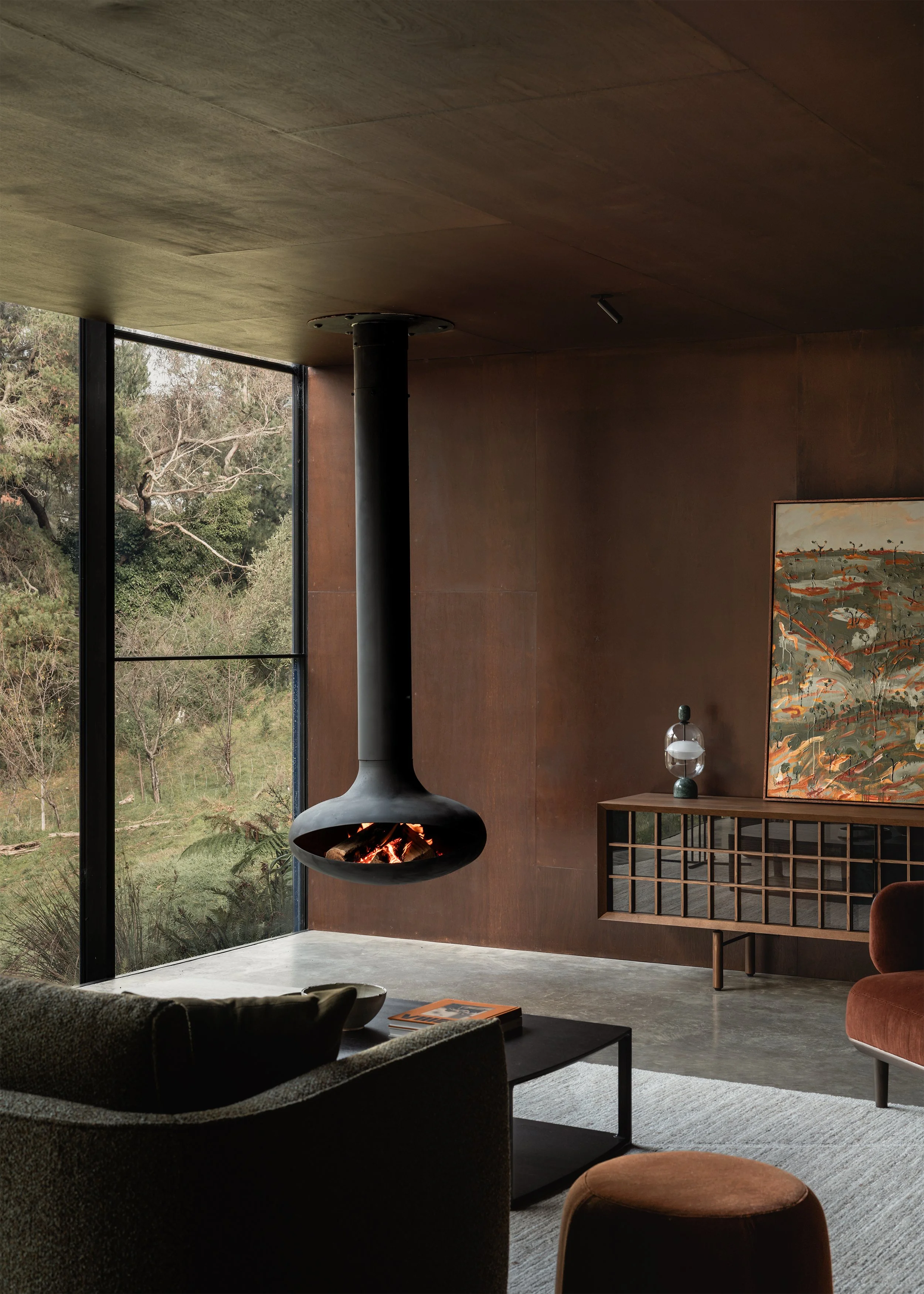 Suspended fireplace and armchair beside a tall bushland window in the dark timber-lined living room.