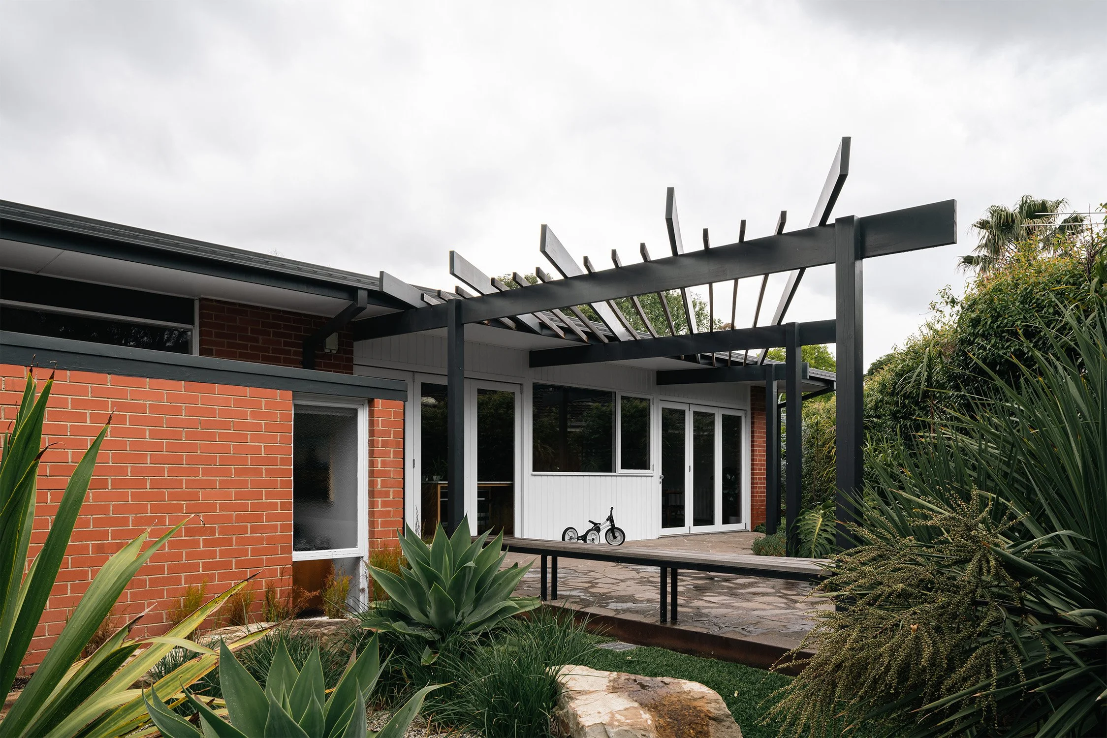 Black pergola over a stone-paved terrace, with a long timber bench and succulent planting at an Australian mid century home.