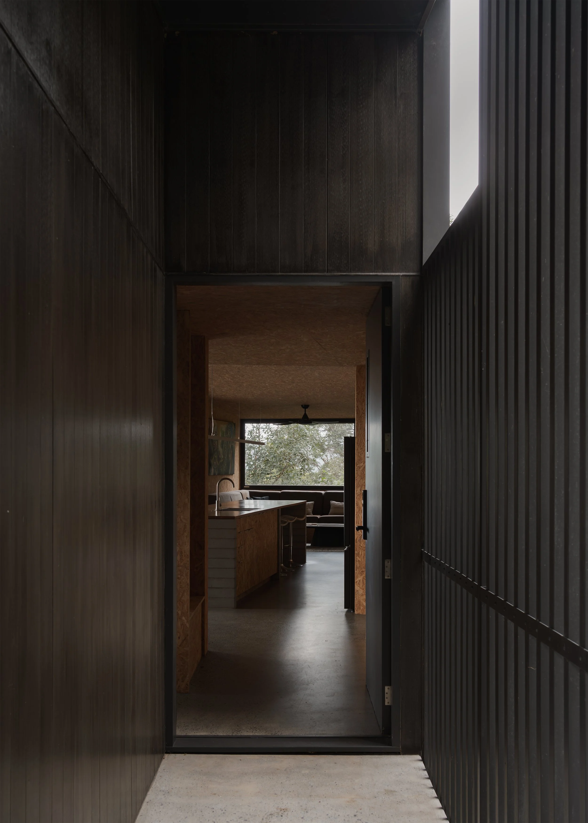 View through the dark entry passage of Tallowwood Cabin into the warm OSB-lined kitchen and living space.