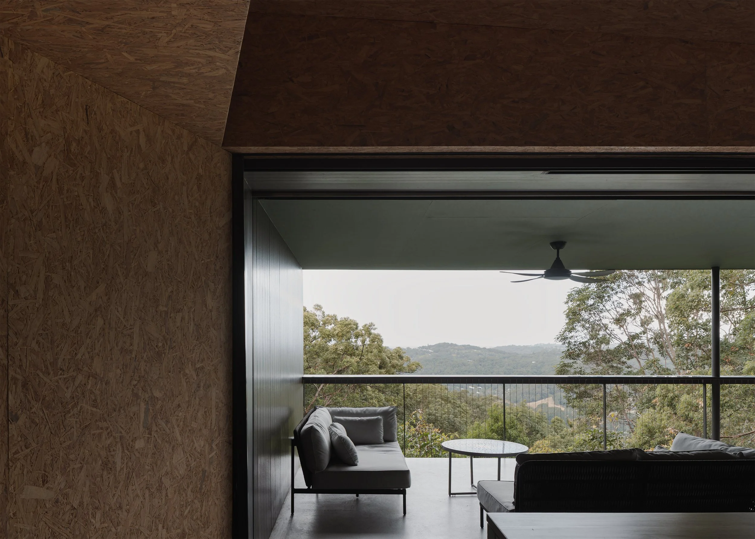View from the OSB-lined interior of Tallowwood Cabin to the covered deck and forested hills of the Gold Coast hinterland.