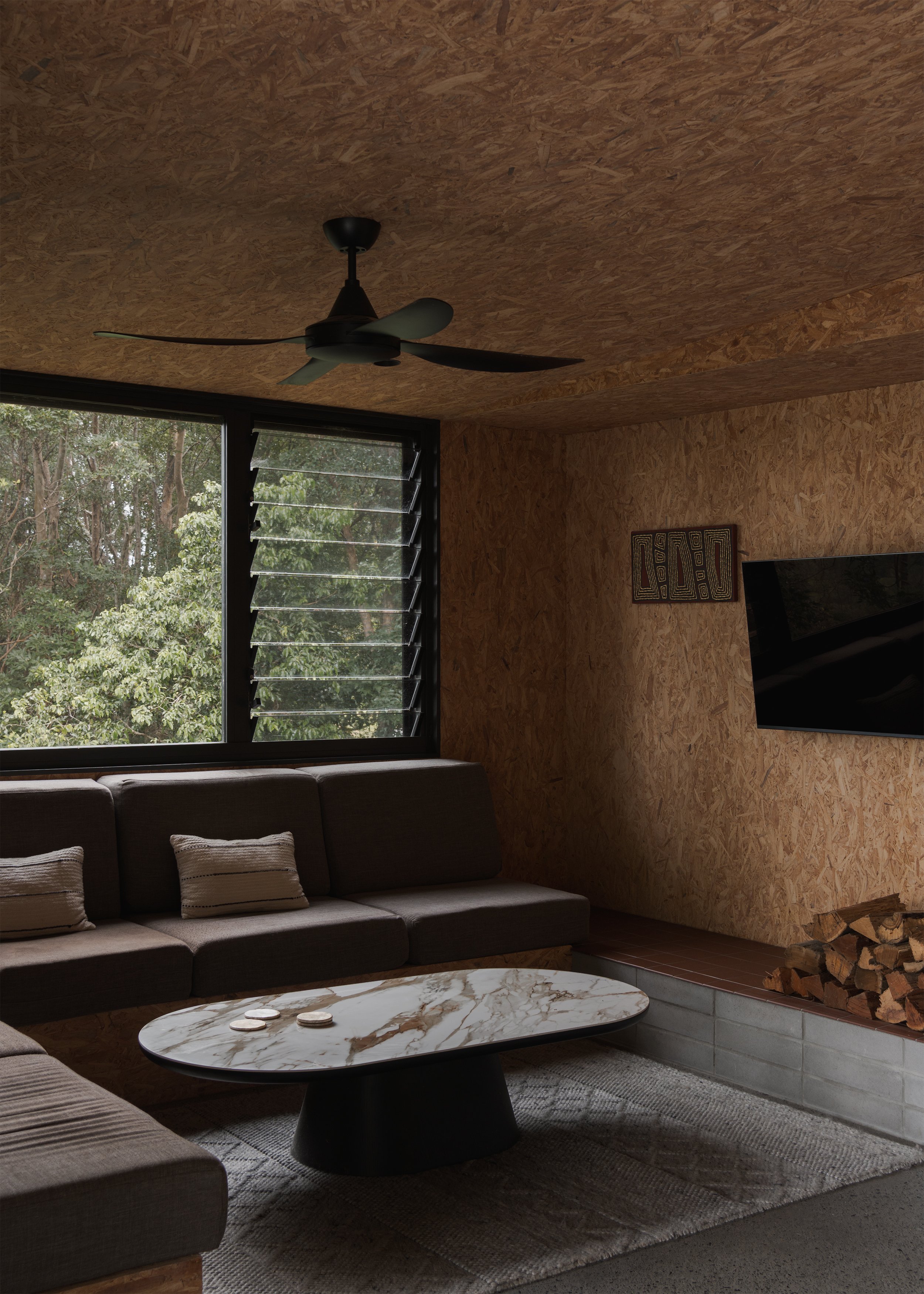 Built-in living area at Tallowwood Cabin with exposed OSB surfaces, a fireplace ledge and broad windows facing the surrounding canopy.