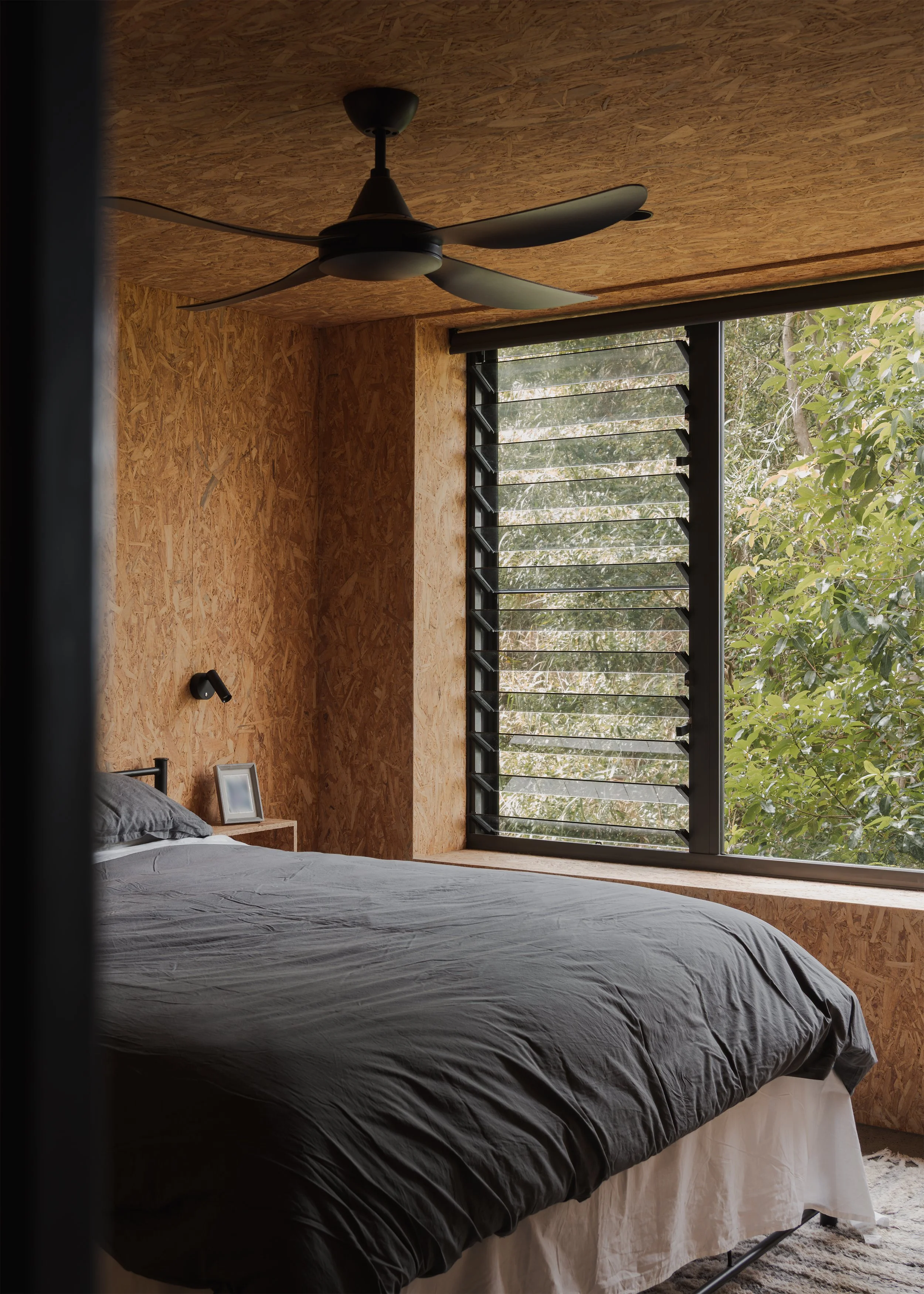 Bedroom at Tallowwood Cabin with exposed OSB walls and ceiling, dark bedding and louvred windows set beside the tree canopy.