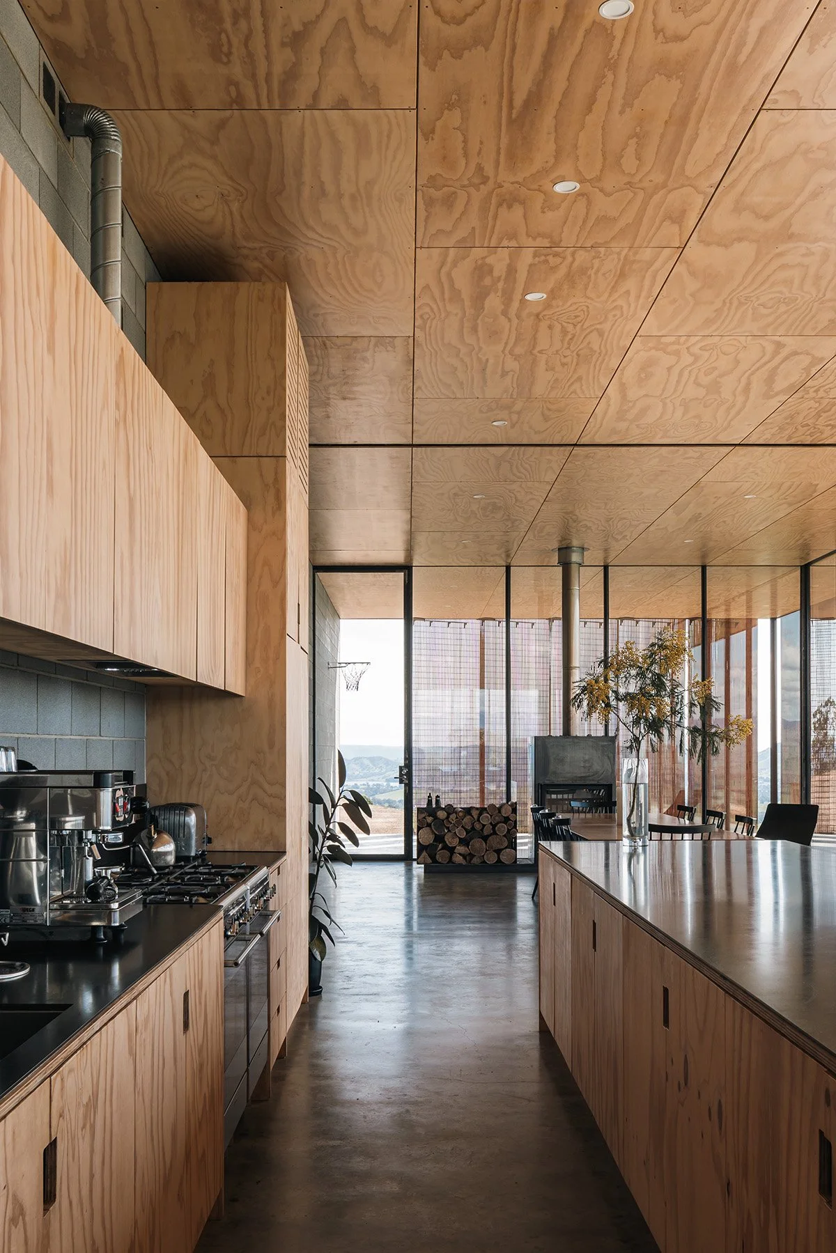 Long plywood kitchen and island beneath a plywood ceiling, looking toward the glazed dining area of Mansfield House.