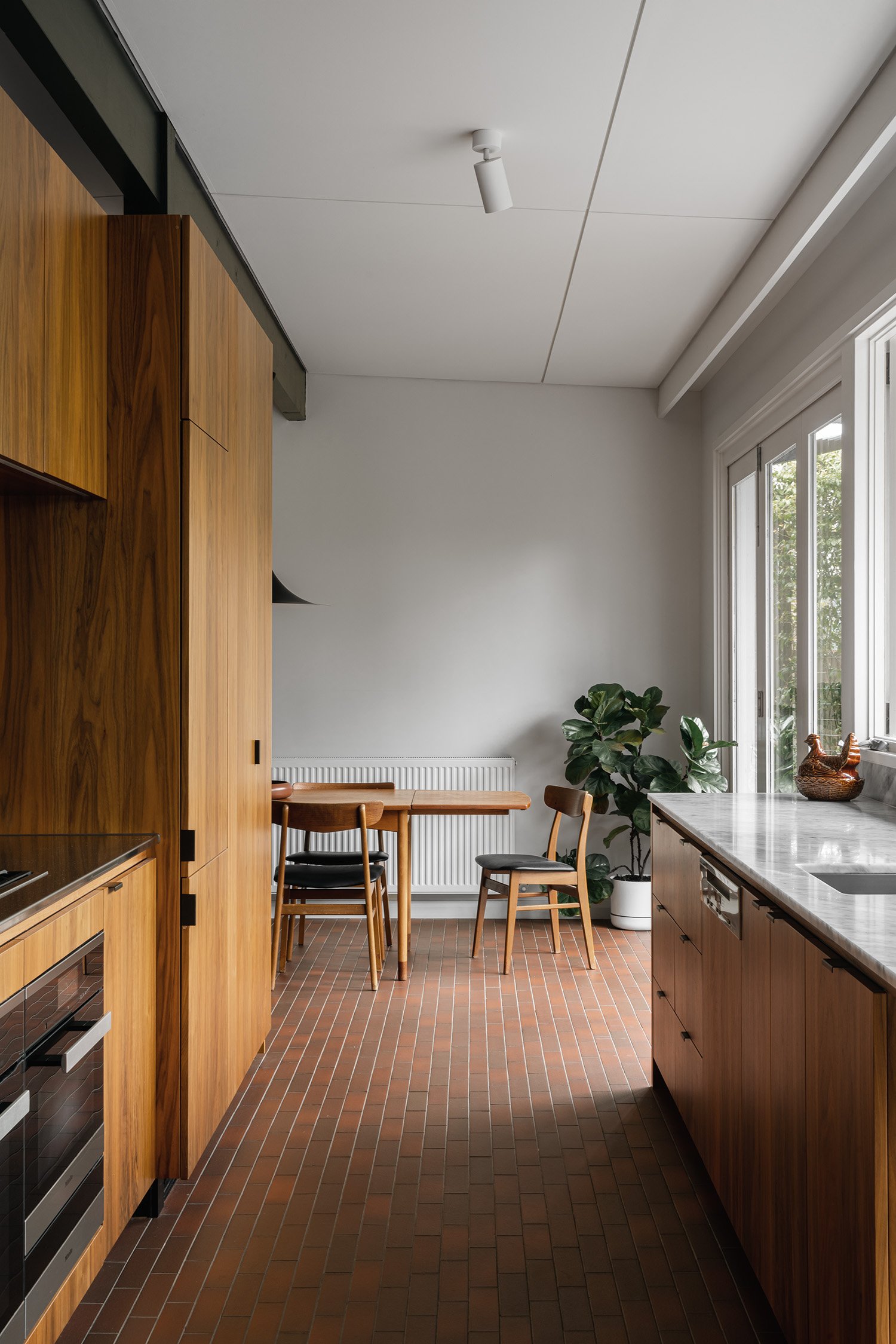 Galley kitchen opening to a timber dining setting in an Australian mid century home, with walnut joinery and terracotta tiles.