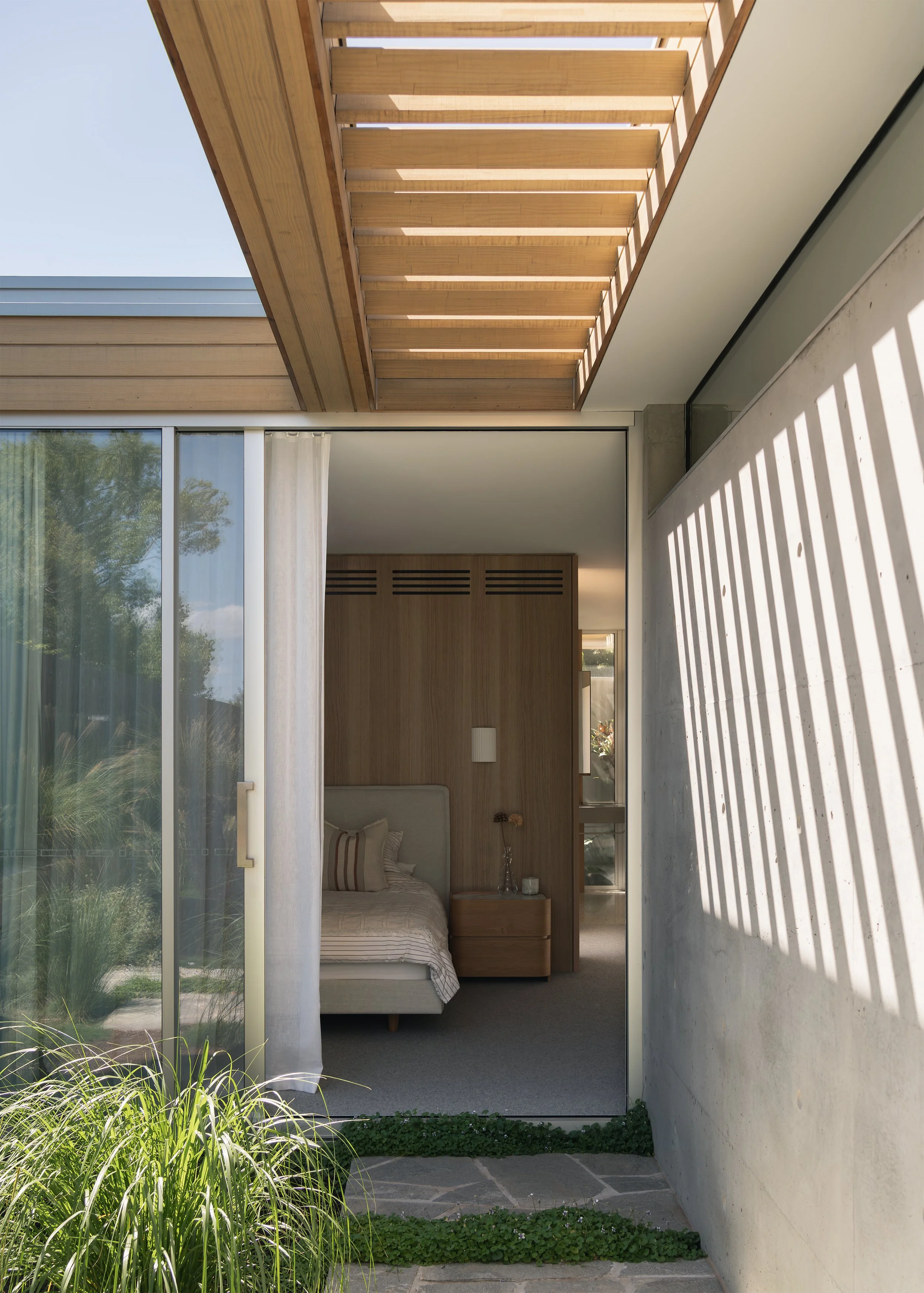 Bedroom opening to a planted garden beneath a timber pergola at Channel by Megowan Architectural in Mornington.