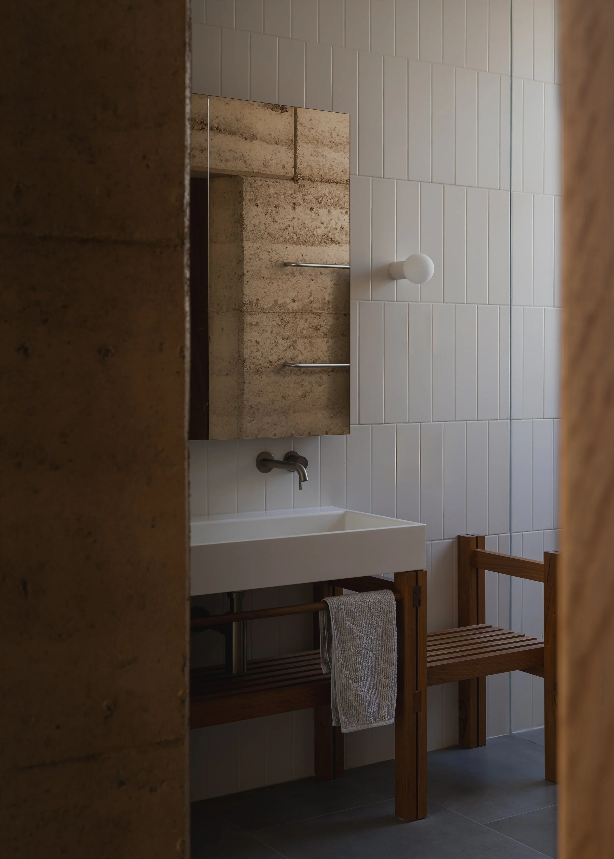 Bathroom at Forest Road House with a white basin, timber vanity frame and vertical white tiles beside an exposed rammed-earth wall.