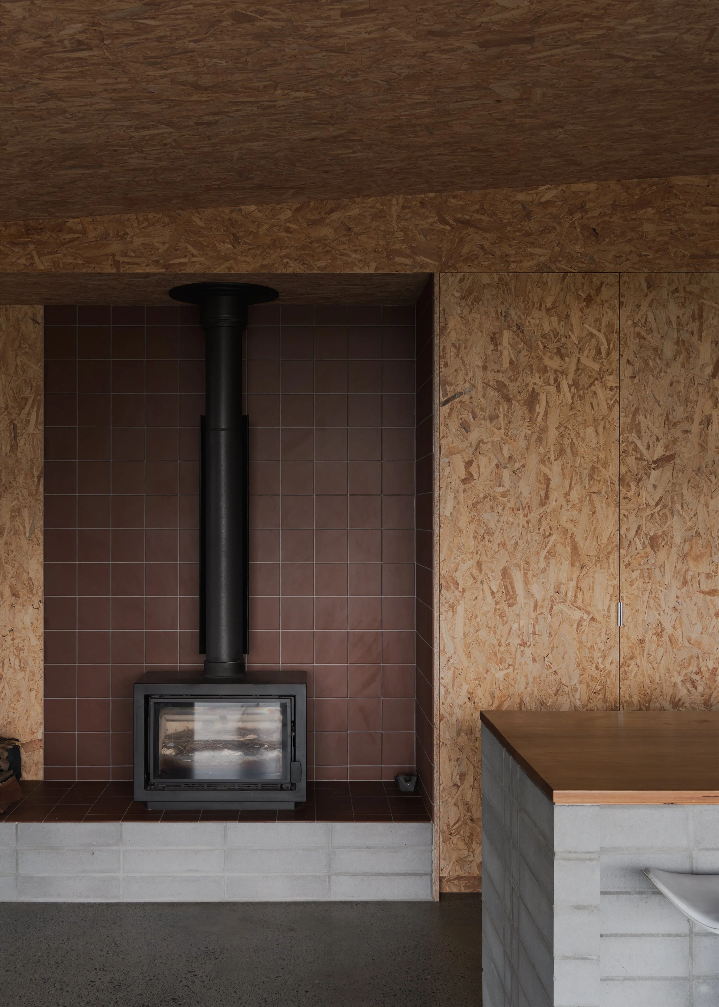 Black wood-burning fireplace set within brown tiles and exposed OSB joinery beside the concrete-block kitchen island at Tallowwood Cabin.