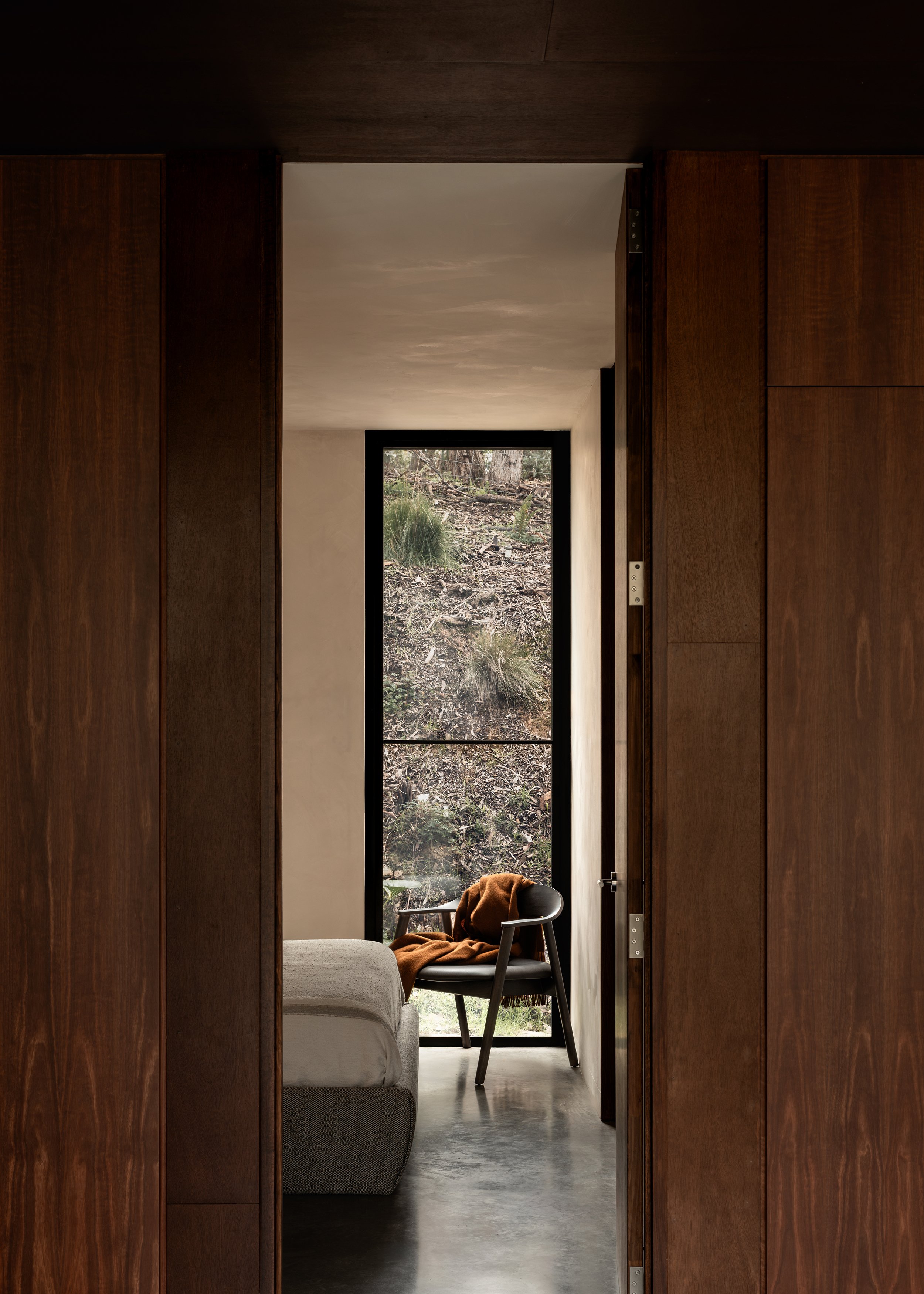 Bedroom framed by dark timber walls, with a narrow full-height window looking directly onto the bushland.