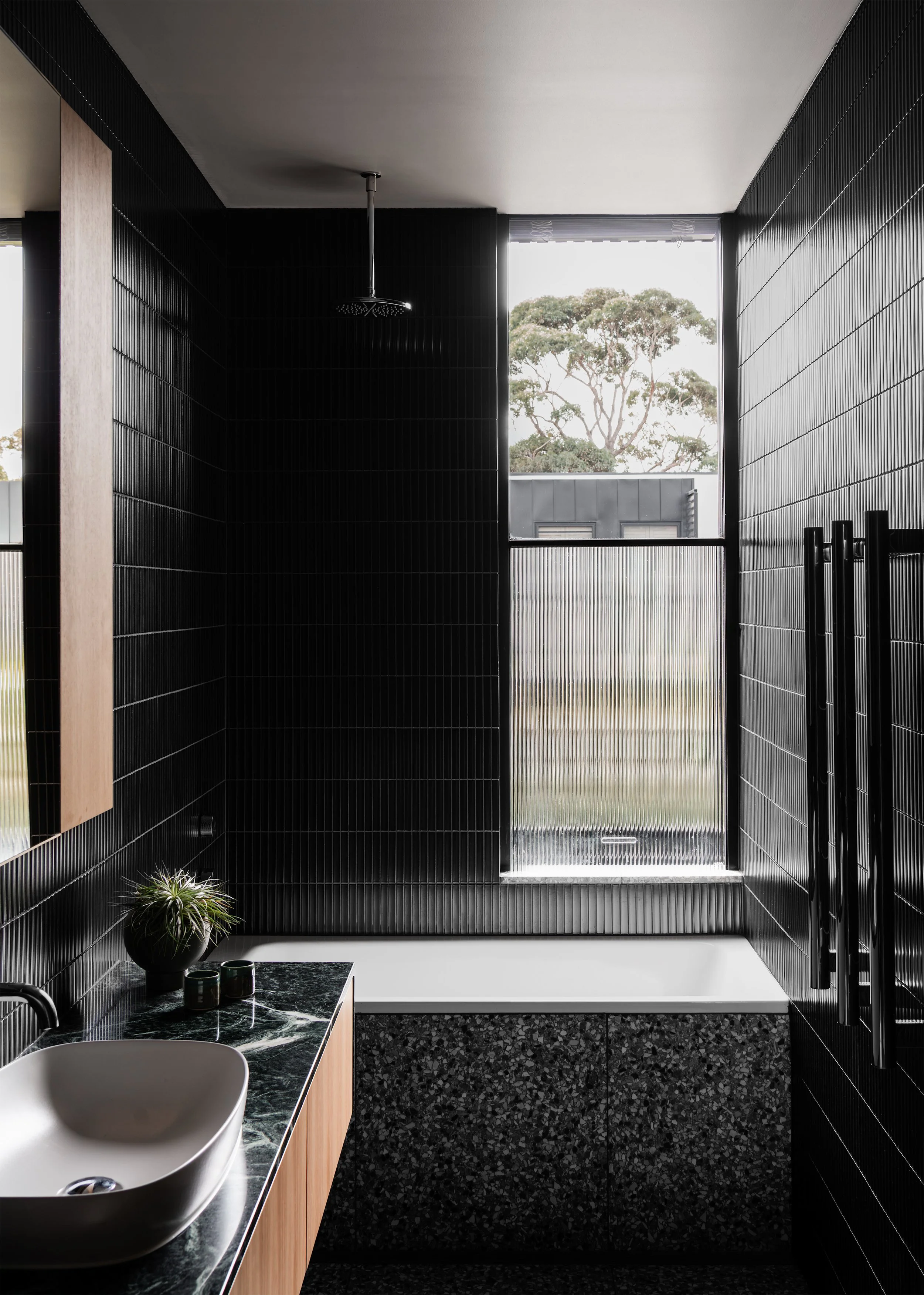 Black-tiled bathroom with bath, stone vanity and framed treetop view above privacy glazing.