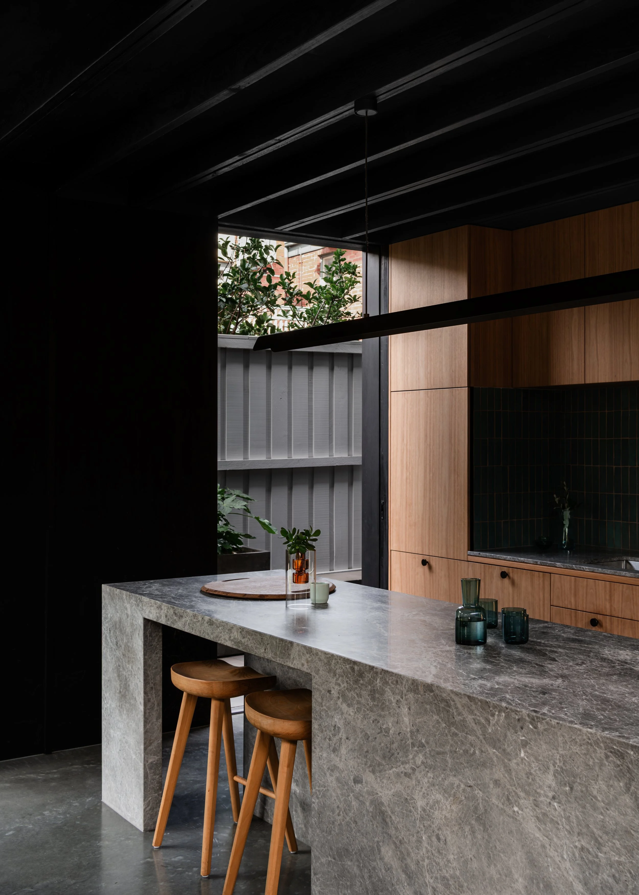 Timber kitchen and stone island beneath the dark roof structure in Topology Studio’s Melbourne extension.