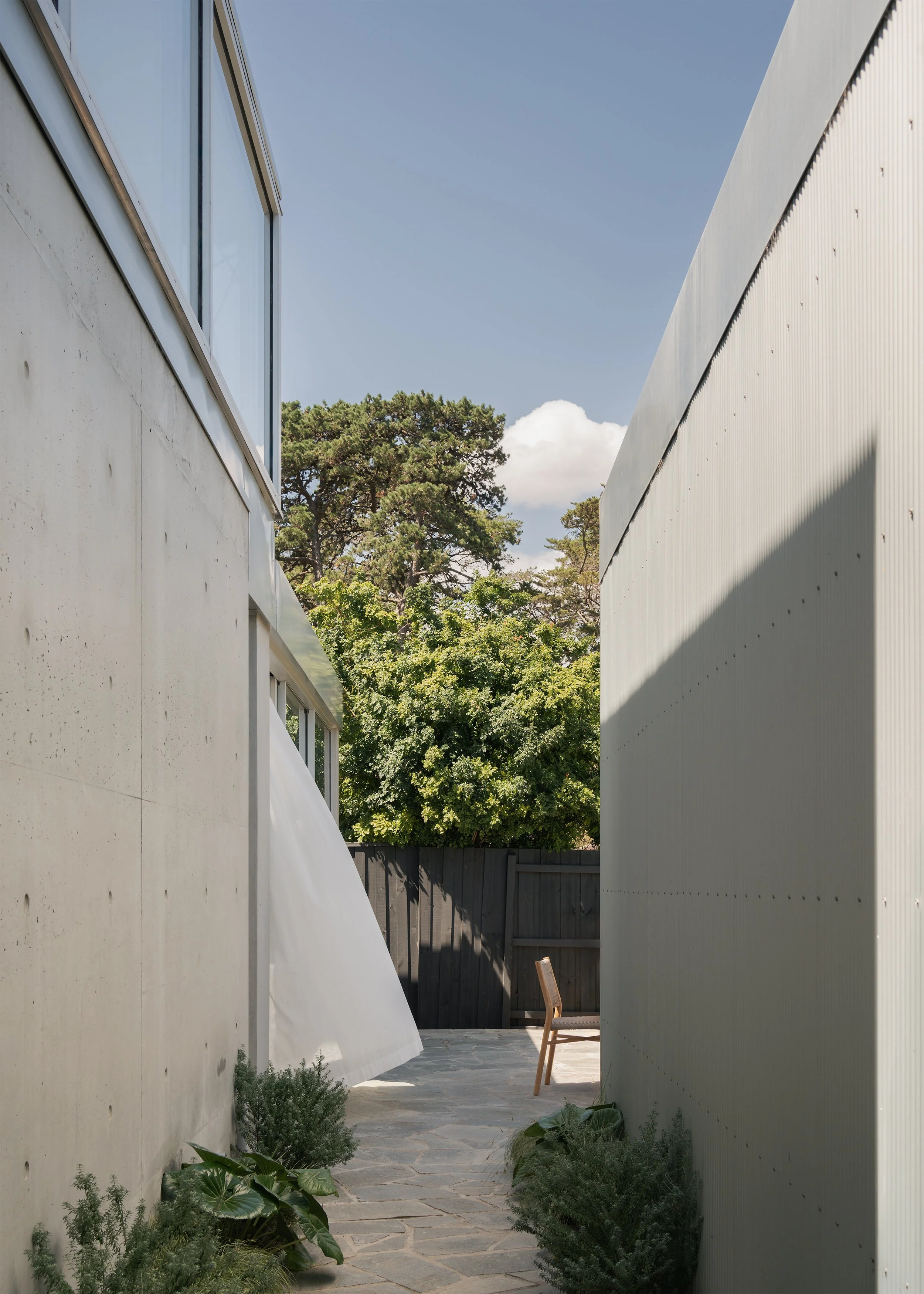 Narrow paved passage between exposed concrete and corrugated walls, framing mature trees beyond Channel.