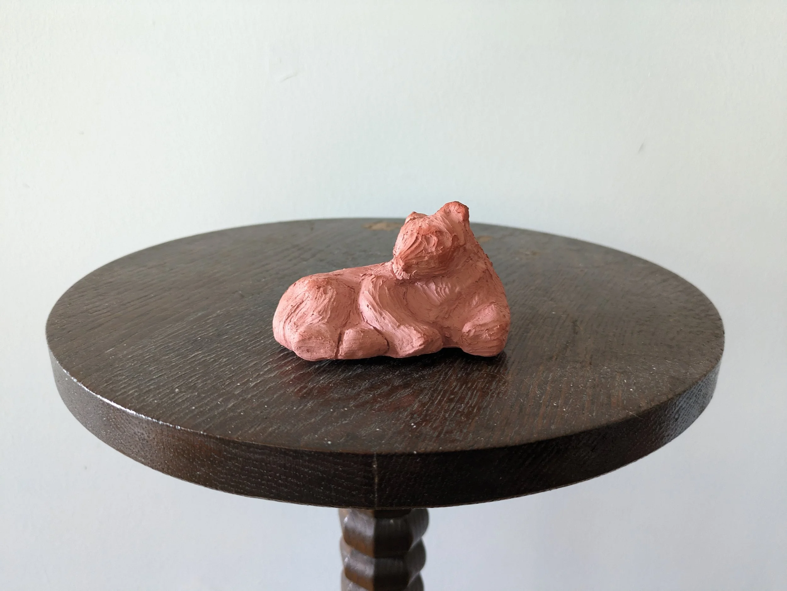 A small, pink, textured sculpture of a reclining lion on a round, dark wooden table against a plain, light-colored wall.