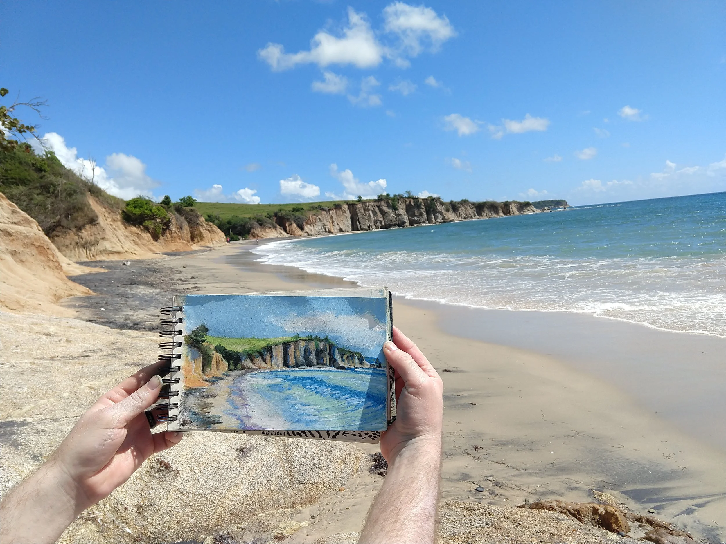 Person holding a sketchbook with a drawing of a beach scene, in front of a real beach with cliffs, sand, waves, and a blue sky with clouds.