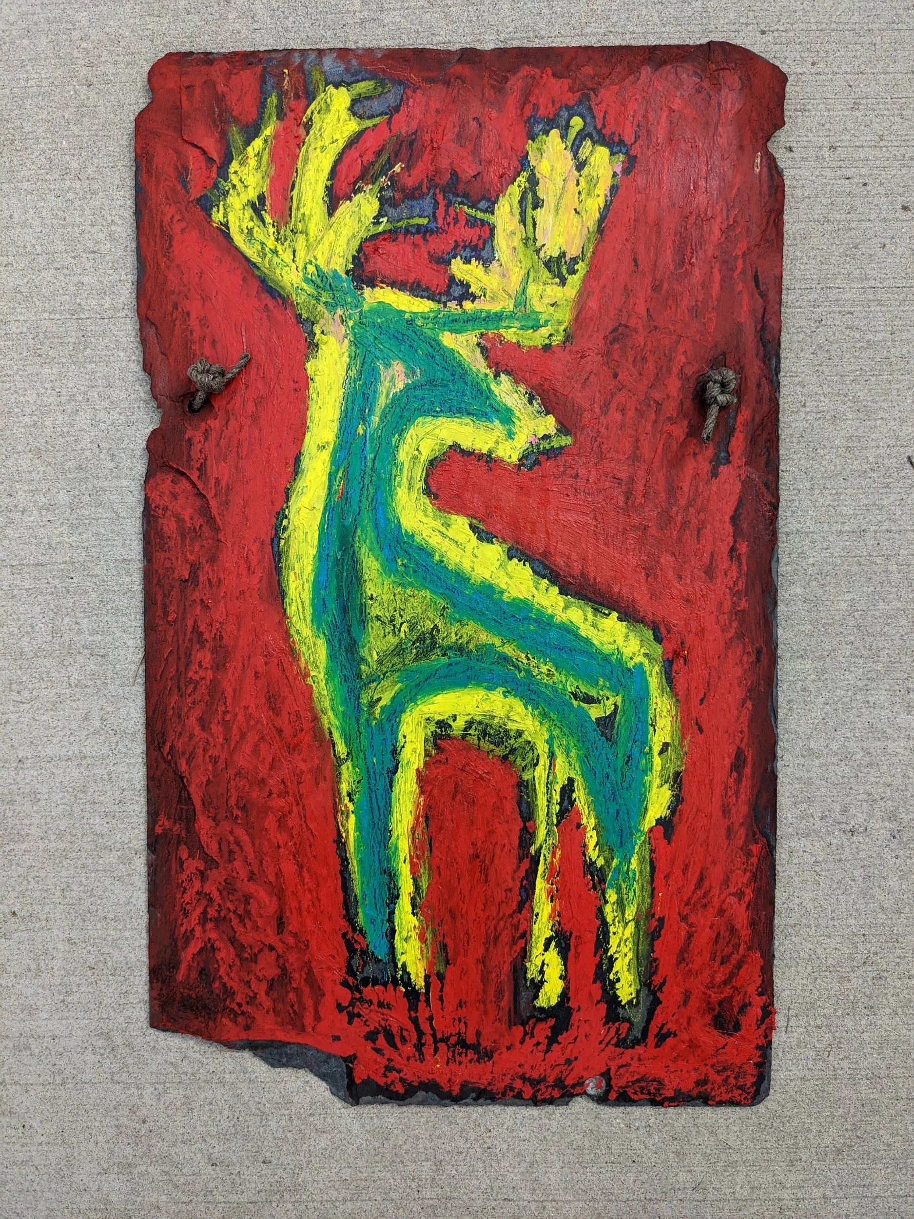 A colorful abstract painting of a reindeer on a rough red background, mounted on a gray wall with two knots attached on sides.