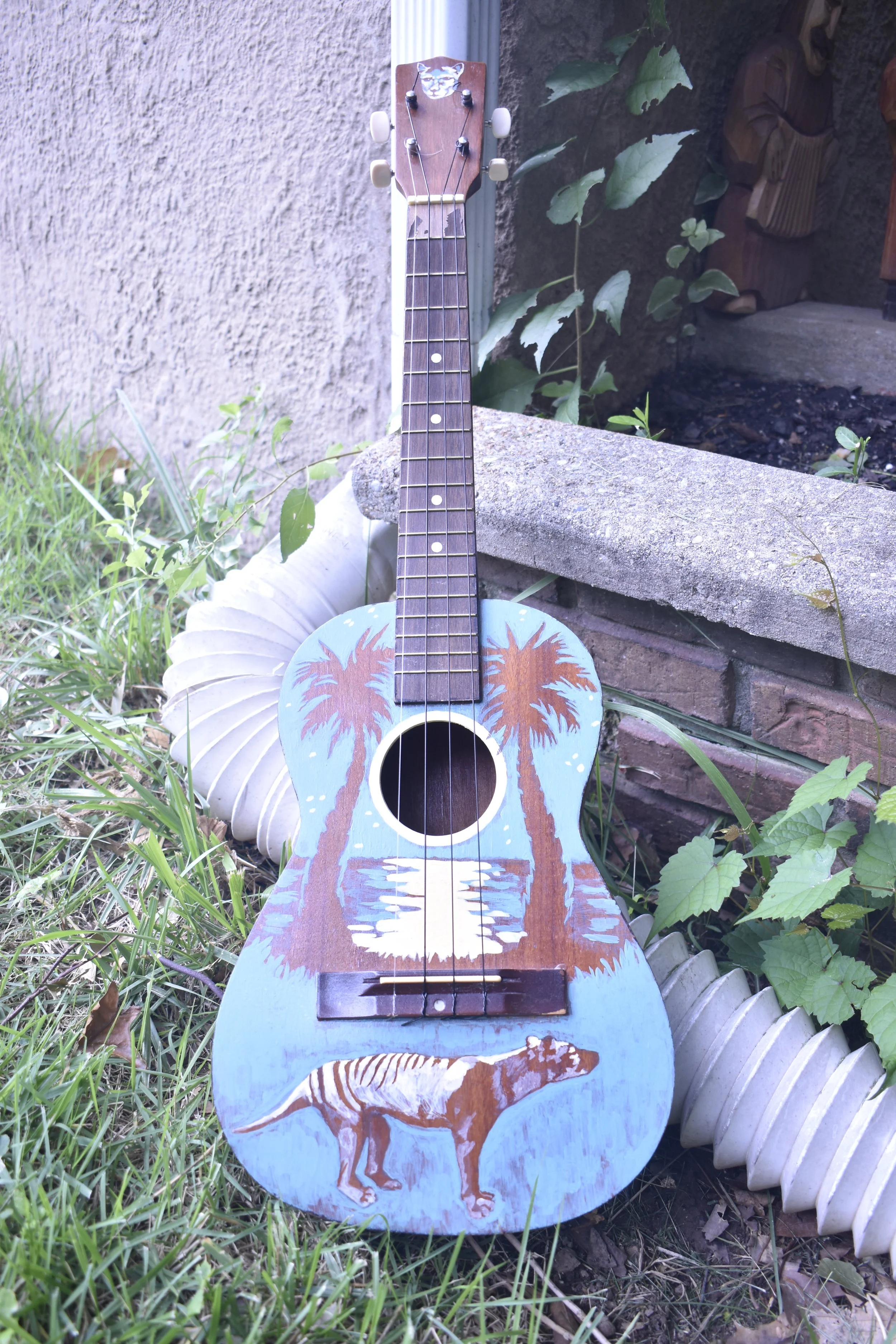 A small acoustic guitar with a hand-painted beach scene, including palm trees, water, and a dinosaur, resting outdoors against grass, a white garden decoration, and a garden wall with greenery.