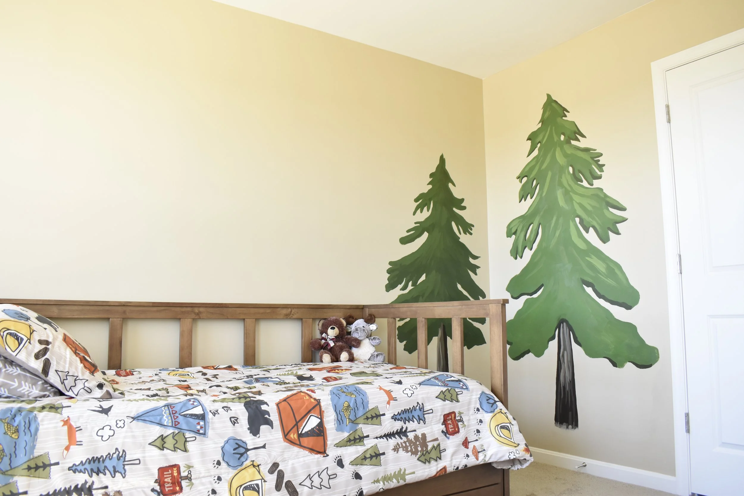 A bedroom with a mural of two green pine trees painted on the wall behind the bed. The bed has a colorful outdoor adventure-themed bedspread with tents, trees, and trails. There are two stuffed bears sitting on the bed near the wall.