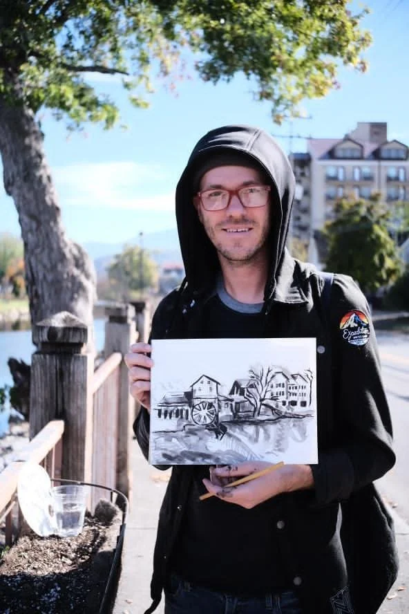A man wearing a hoodie and glasses holding a drawing of a landscape with buildings, trees, and a river, standing outdoors near a wooden railing with greenery and buildings in the background.