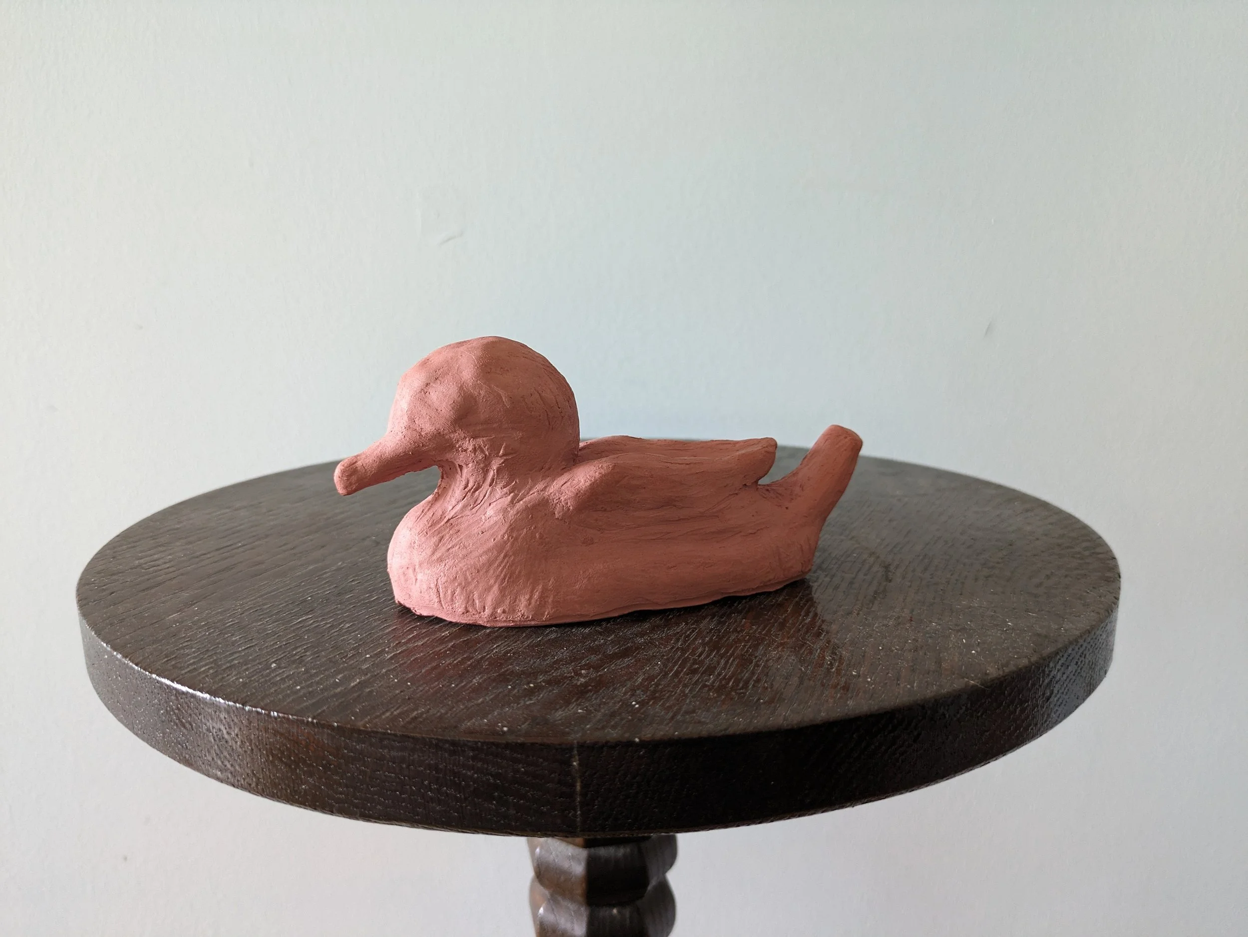 A small, pinkish-brown duck figurine made of a textured material, placed on a dark wooden round table against a plain, light-colored wall.