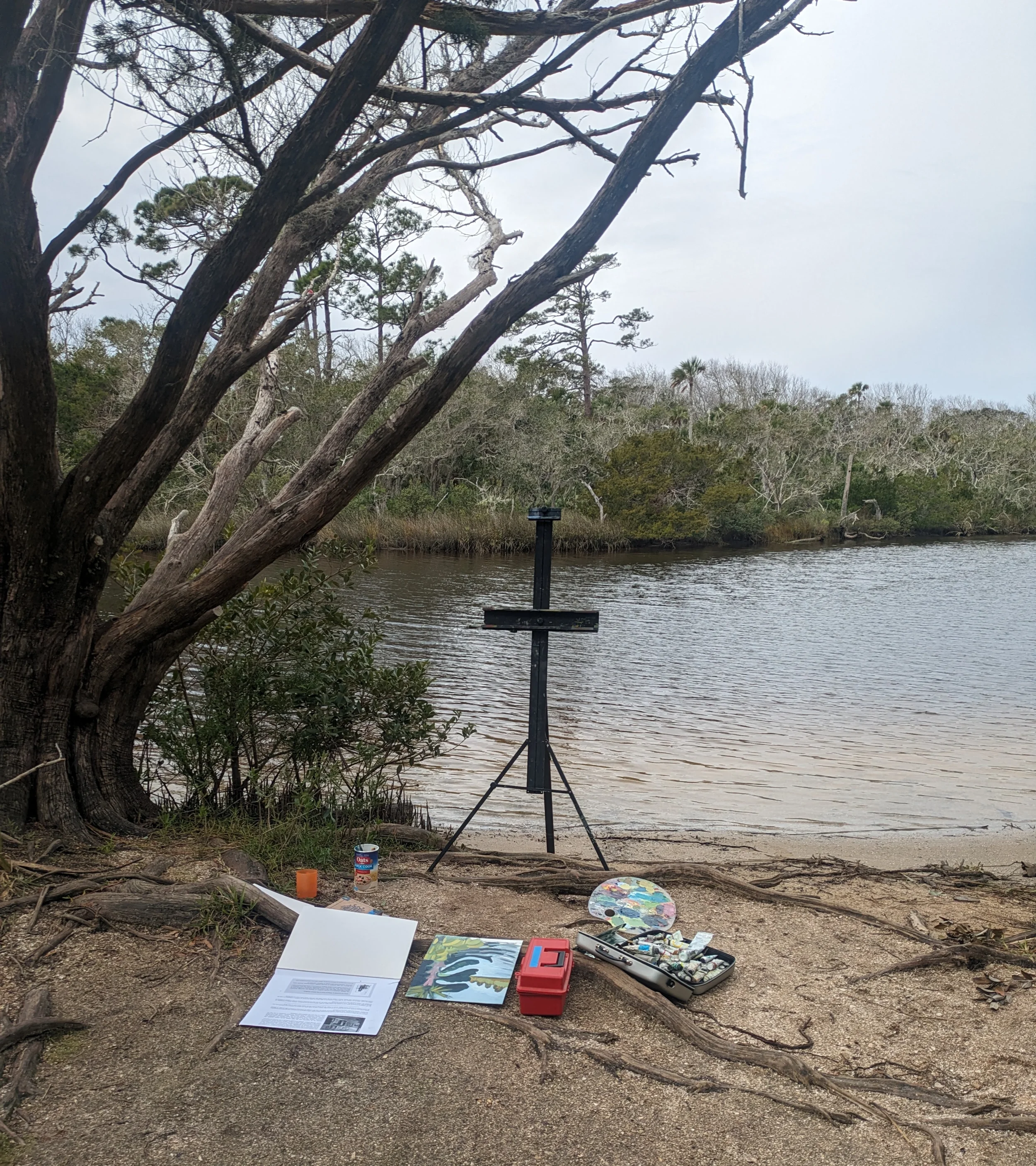 An outdoor painting setup by a lakeside with trees, water, and cloudy sky. Art supplies, a palette, paint tubes, brushes, and a red toolbox are on the ground. An easel stands ready for painting.