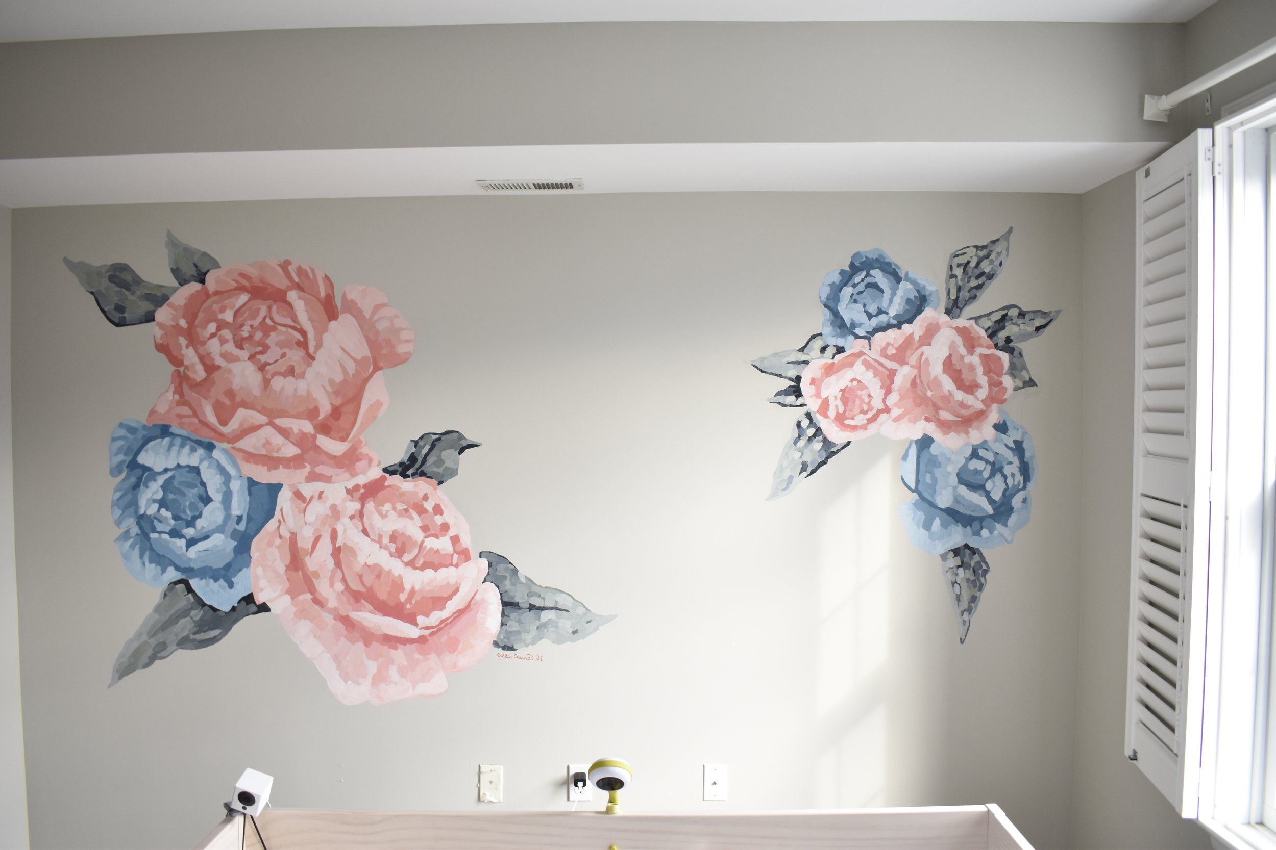 Wall with large floral mural of pink and blue roses with gray leaves in a bright room with a window and white shutter.