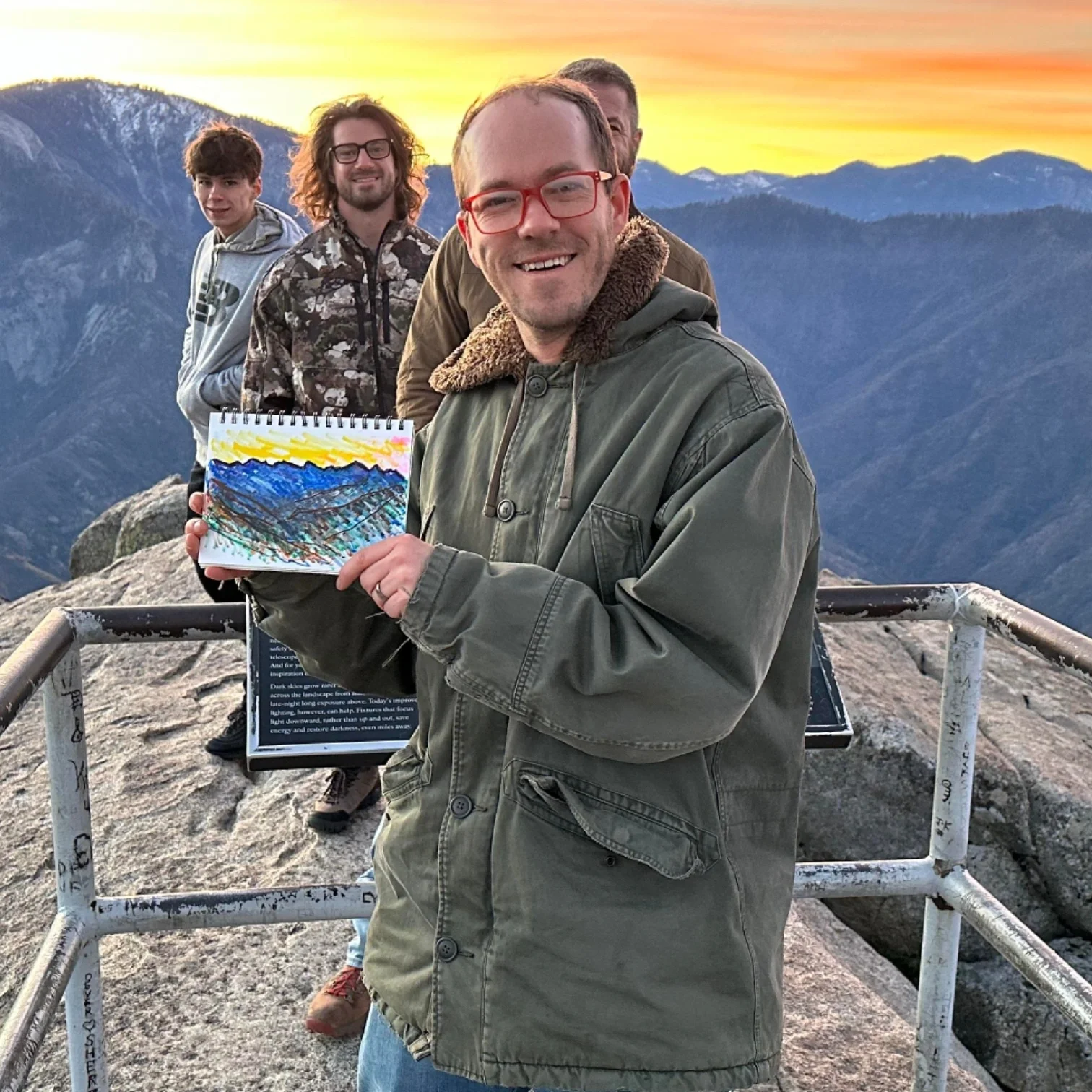 Four men standing on a mountain overlook at sunset, with mountains in the background. The man in front is smiling and holding a small colorful painting of the mountain landscape.