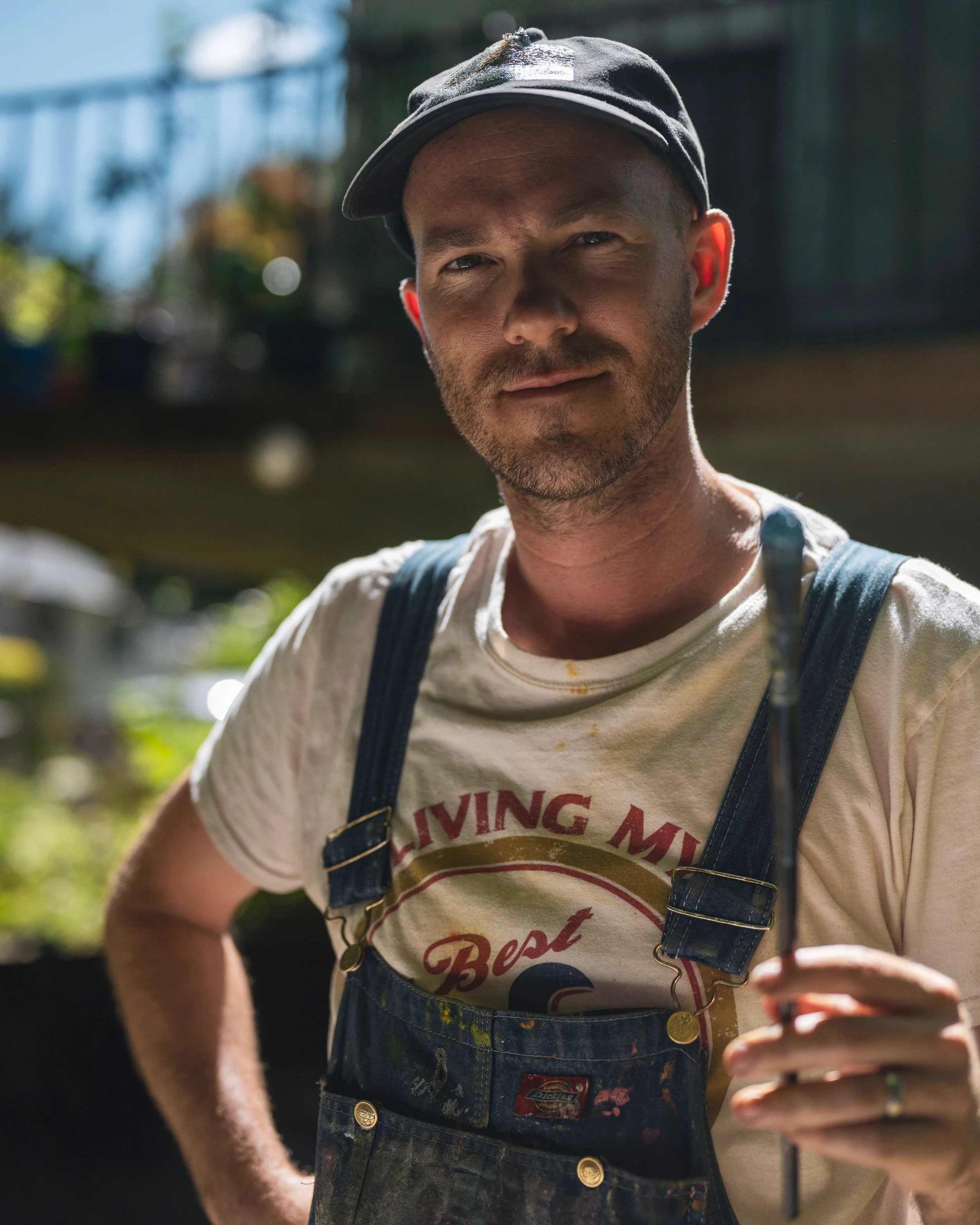 A man wearing a dark baseball cap, a white t-shirt with red and black lettering, and blue overalls holding a gardening tool outdoors with a blurred background.