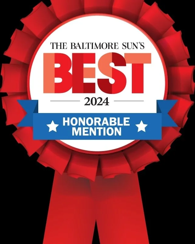 Award ribbon with red and white colors, gold accents, and a blue banner that reads "Honorable Mention" in white text. The center text says "The Baltimore Sun's Best 2024."