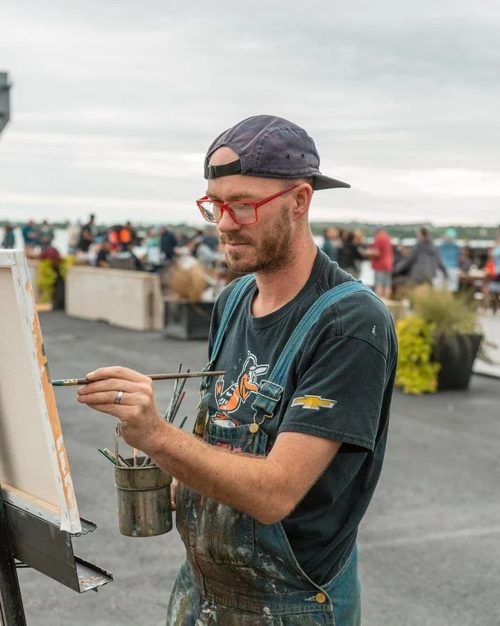An artist wearing a backwards cap, red glasses, and a black t-shirt with a NASCAR logo, painting on a canvas outdoors with a crowd in the background.