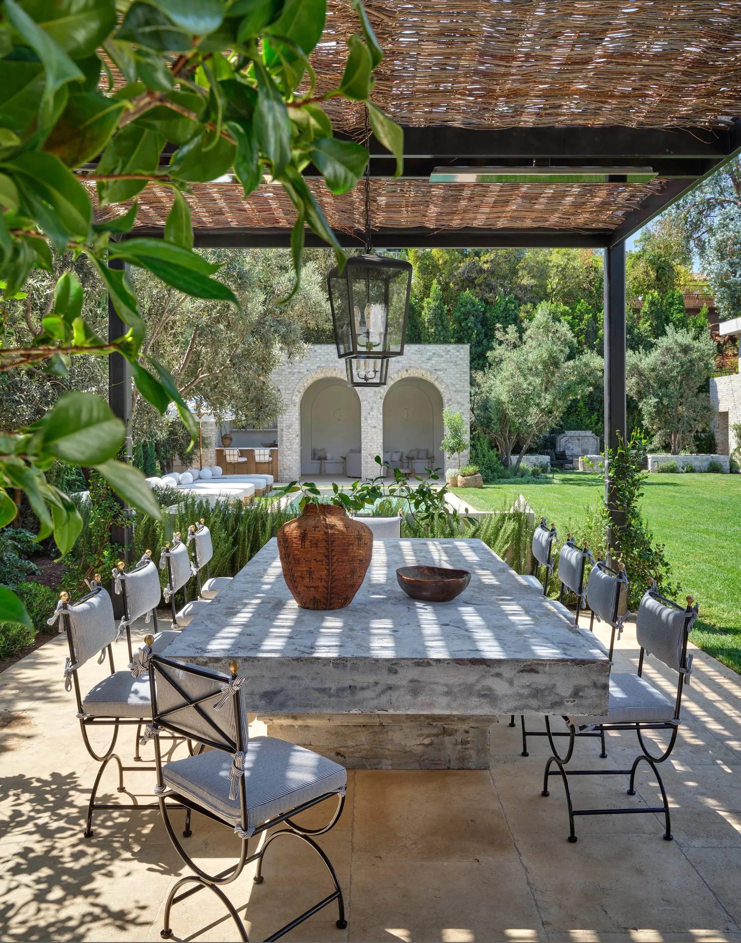 Outdoor dining area with a large stone table, surrounded by cushioned chairs. The area is covered by a pergola with a woven roof. There's potted greenery and a lantern hanging above the table. In the background, a landscaped lawn and arches of a ston