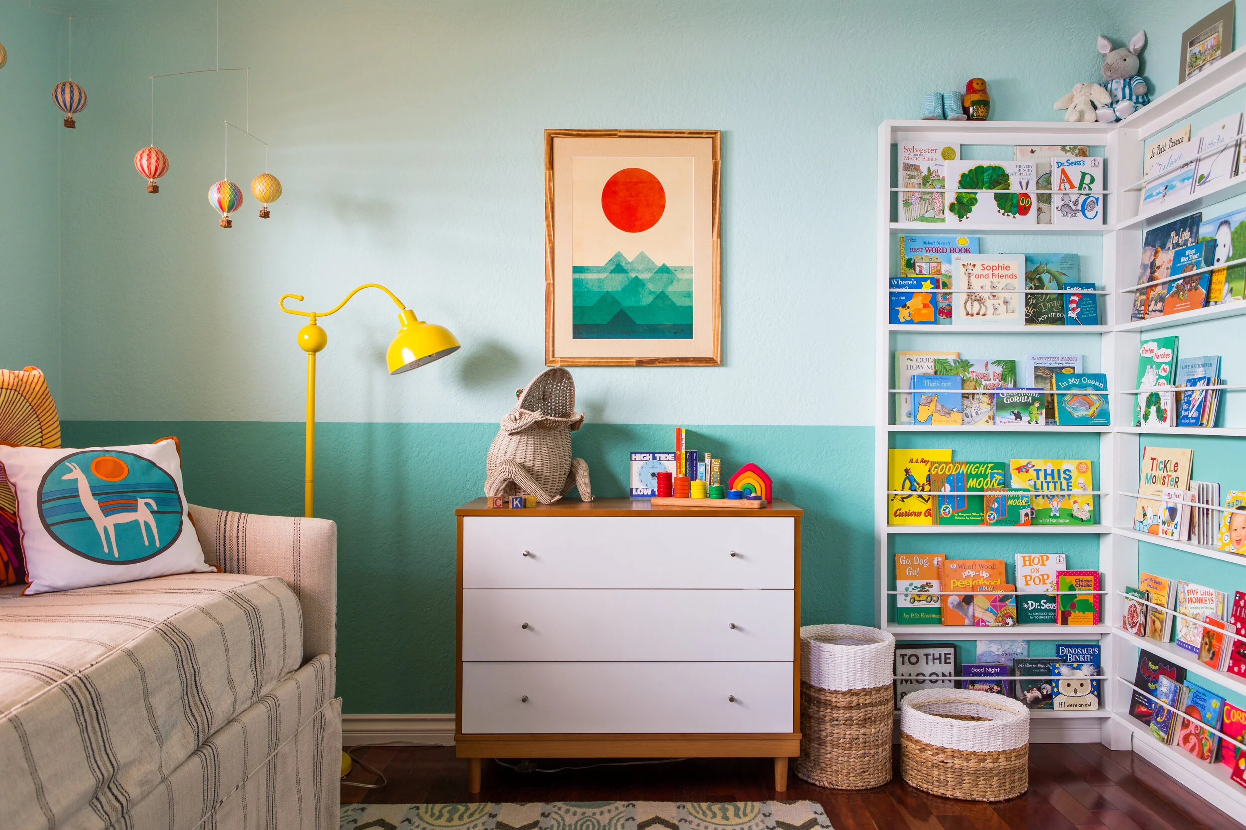 Colorful children's room with a light blue and teal two-tone wall, featuring a dresser with a framed artwork above it. A yellow floor lamp stands next to a bed with a decorative pillow. A mobile of hot air balloons hangs from the ceiling. A bookshelf