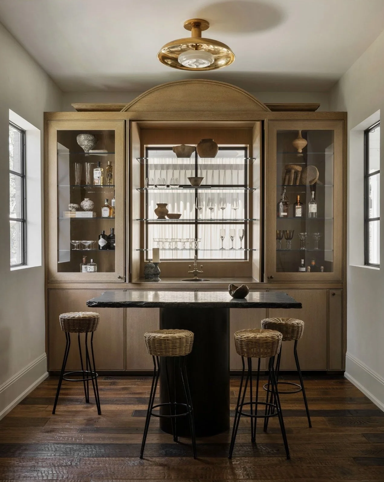 Elegant home mini bar area with wooden cabinetry, glass shelves displaying glassware and bottles, a black stone countertop bar table, and four wicker stools on dark wood flooring. Modern gold ceiling light fixture above.