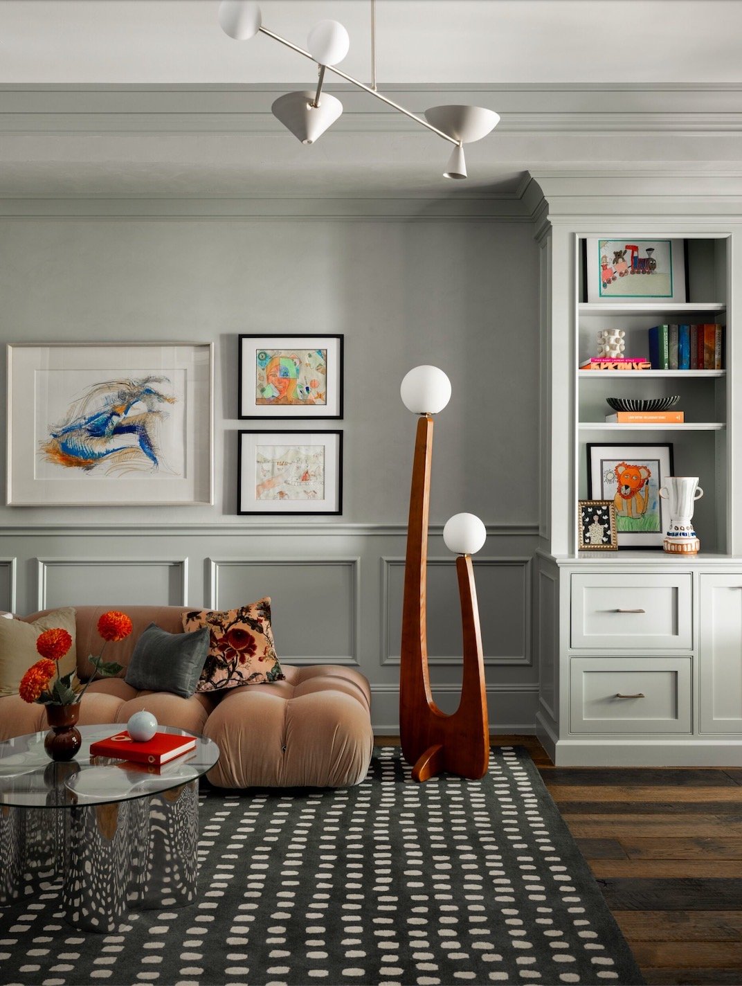 Modern living room with a tufted blush sofa, colorful cushions, abstract wall art, polka dot rug, and a sleek lamp. A metallic coffee table holds red flowers and books. Built-in shelves contain books and decorative items.