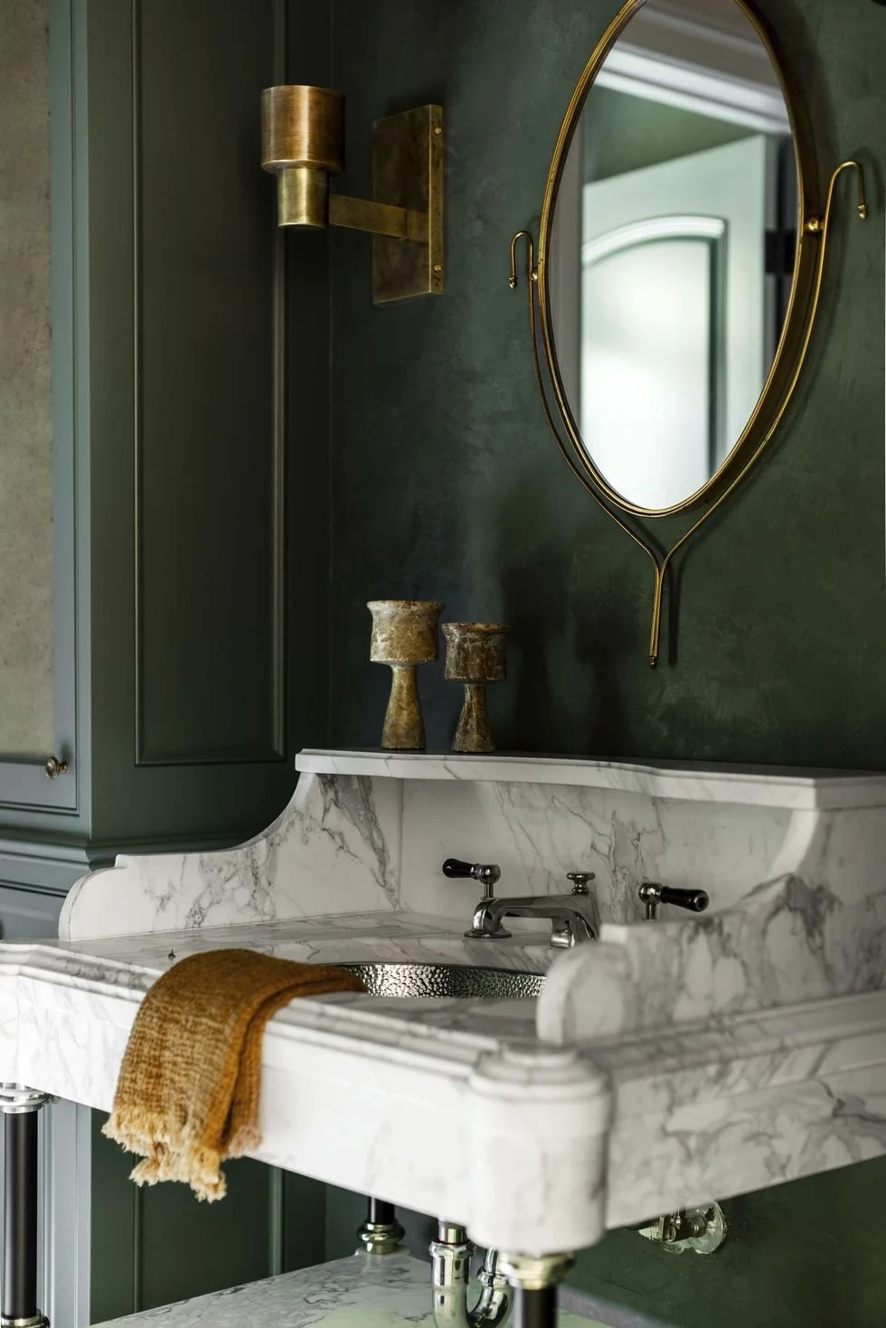 Elegant bathroom with marble sink, green walls, gold oval mirror, and wall-mounted light fixture.